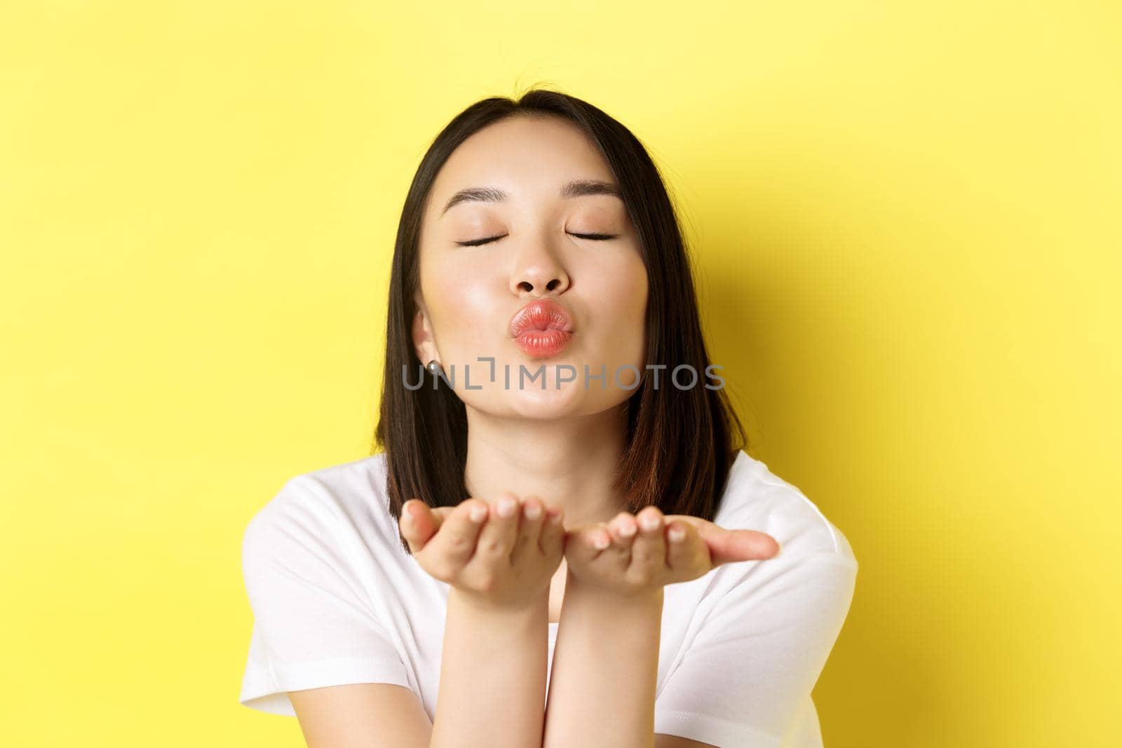 Valentines day concept. Close up of beautiful asian woman pucker lips and holding hands near mouth, blow air kiss at camera with closed eyes, standing over yellow background by Benzoix