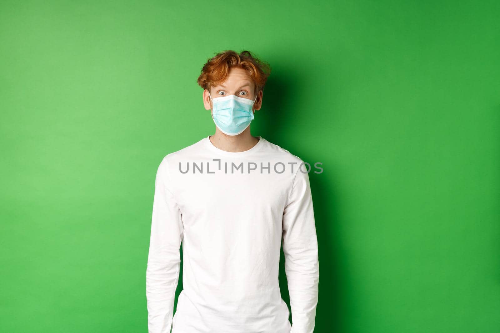 Covid-19, social distancing and lifestyle concept. Young redhead man wearing face mask during coronavirus pandemic, looking surprised at camera, green background by Benzoix