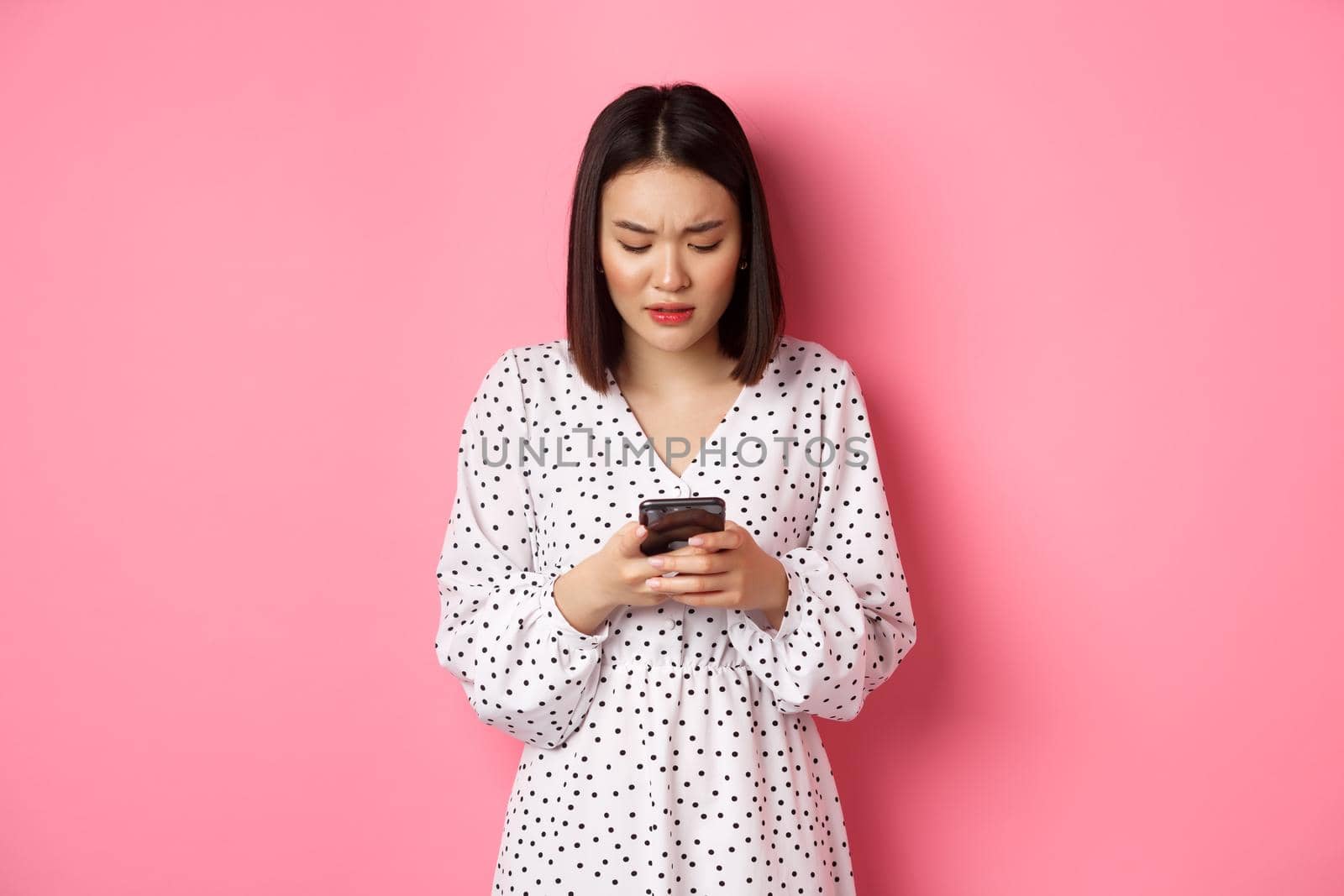 Nervous and concerned asian woman reading message on smartphone, looking worried, standing in dress over pink background.