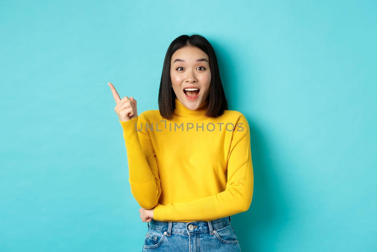 Shopping concept. Attractive korean woman smiling amazed, pointing finger left, showing good deal banner, standing against blue background.