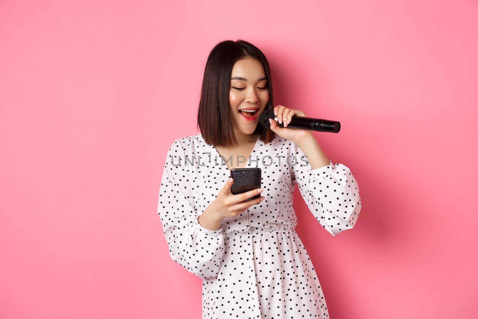 Cute asian woman reading lyrics in smartphone, singing with microphone, standing in trendy dress over pink background.