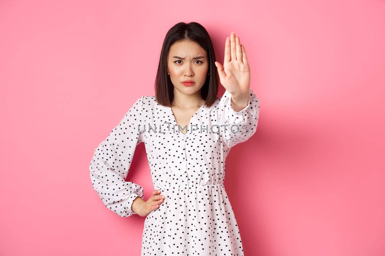 Angry asian woman tell stop, extend arm to prohibit or disapprove something, frowning displeased, standing over pink background.