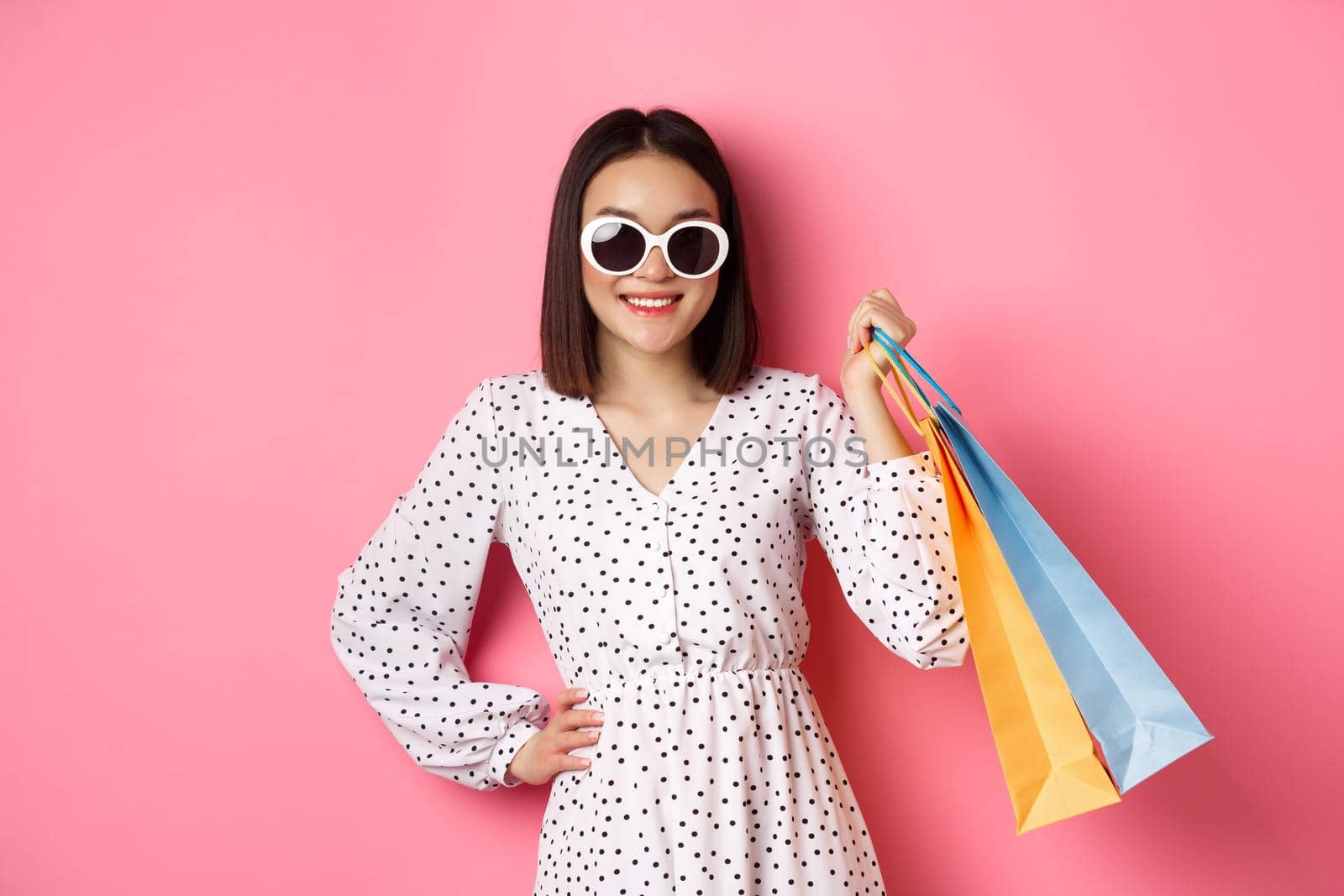 Attractive smiling asian woman holding shopping bags, wearing sunglasses and cute dress, standing against pink background.