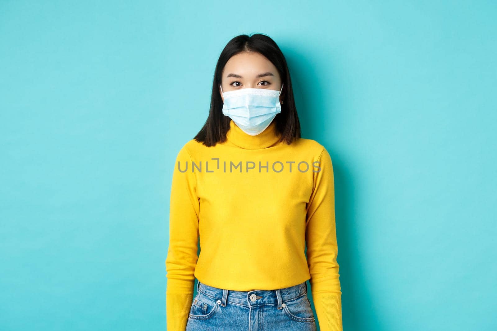 Covid-19, social distancing and pandemic concept. Teenage asian woman in medical mask, looking at camera and standing against blue background in yellow sweater by Benzoix