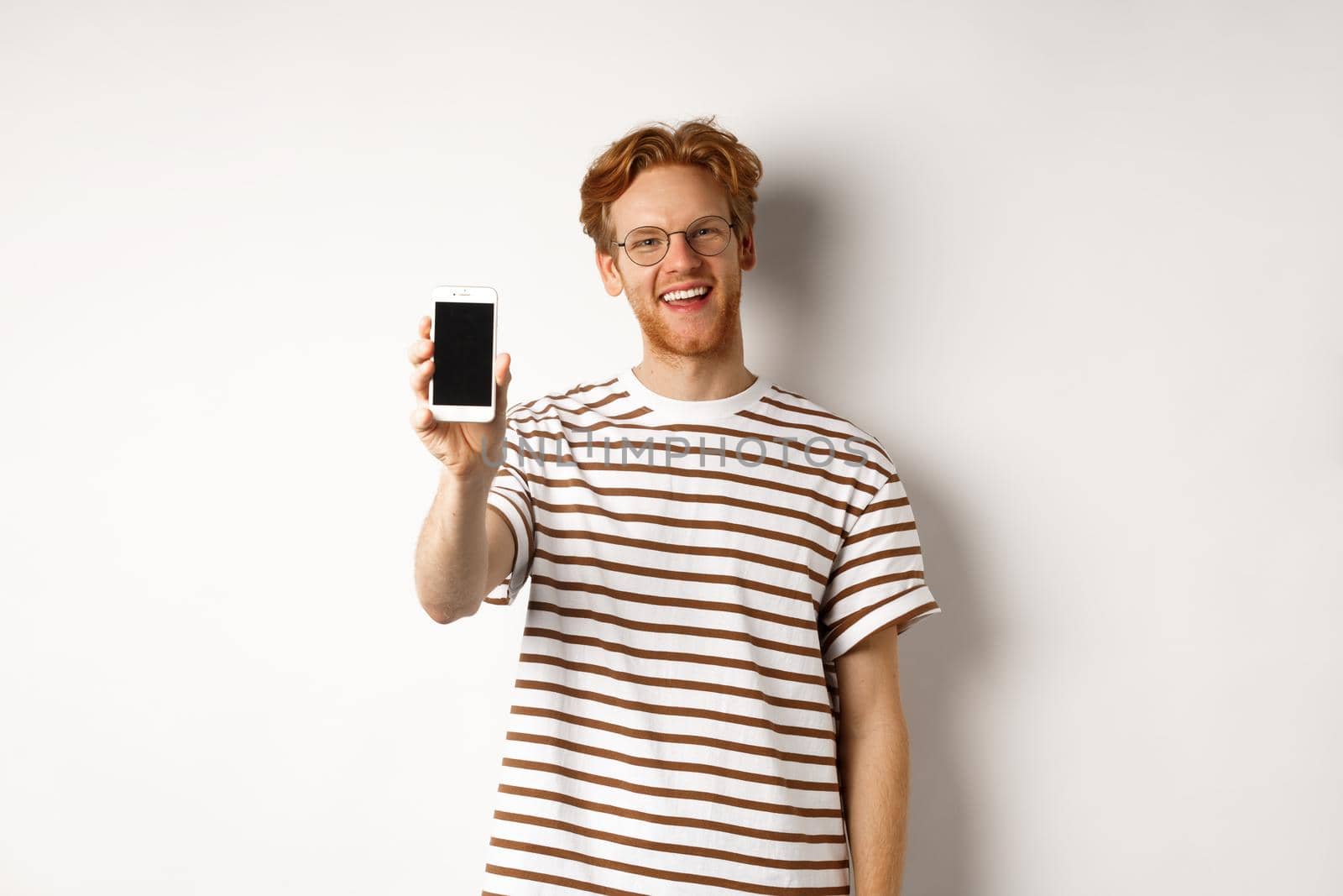 Technology and e-commerce concept. Happy young redhead man in glasses showing blank smartphone screen, smiling satisfied, standing over white background by Benzoix