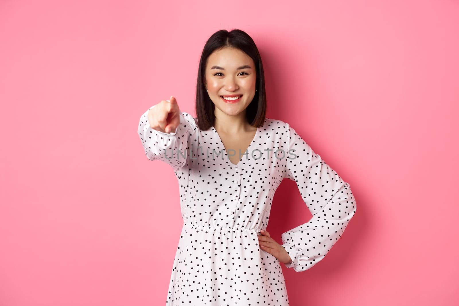 Confident asian woman in dress pointing at you, smiling and staring at camera, standing against pink background by Benzoix