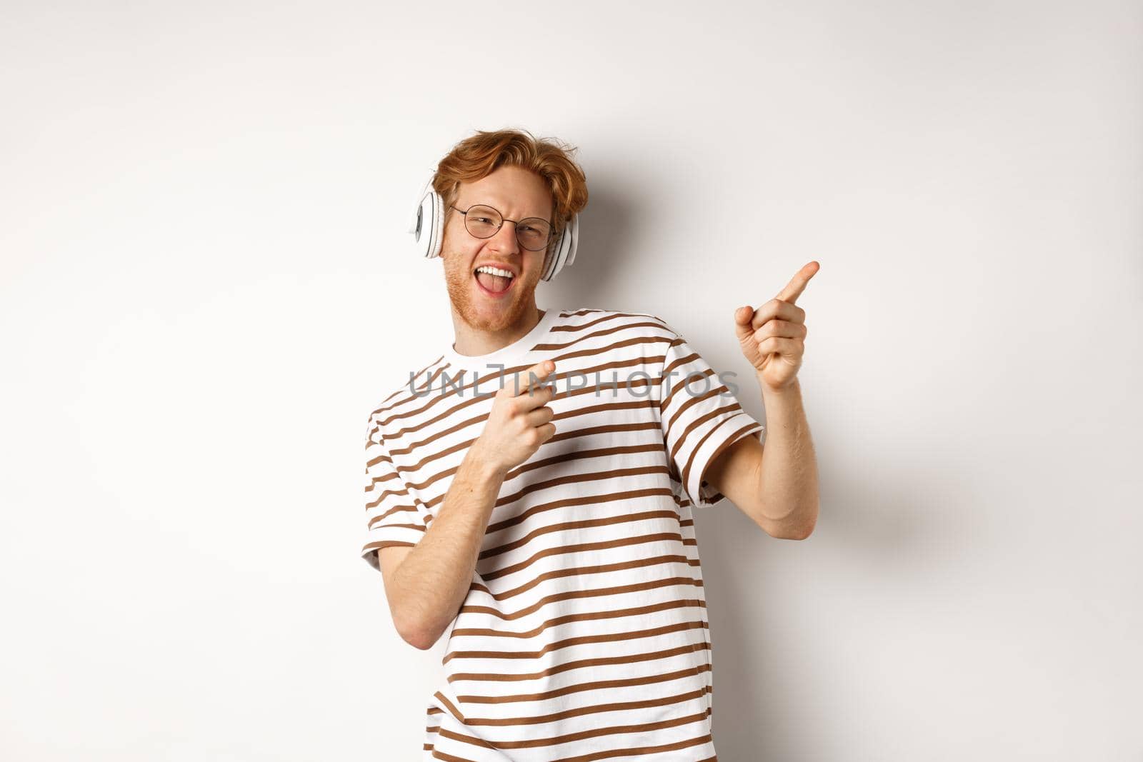 Technology concept. Happy redhead man listening music in headphones and dancing and pointing fingers right, standing over white background.