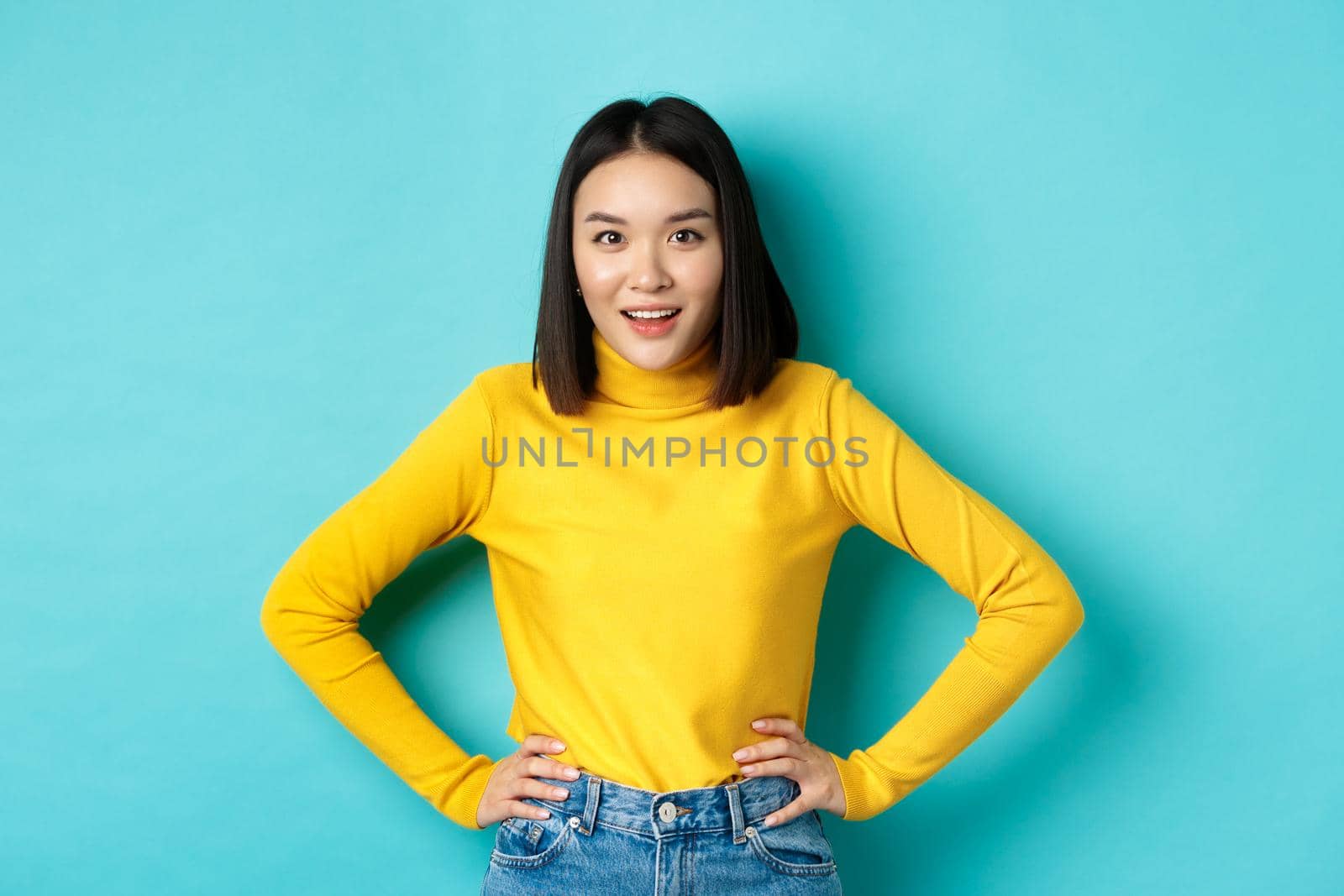 Image of impressed korean girl listening interesting story, looking at camera intrigued, standing over blue background.