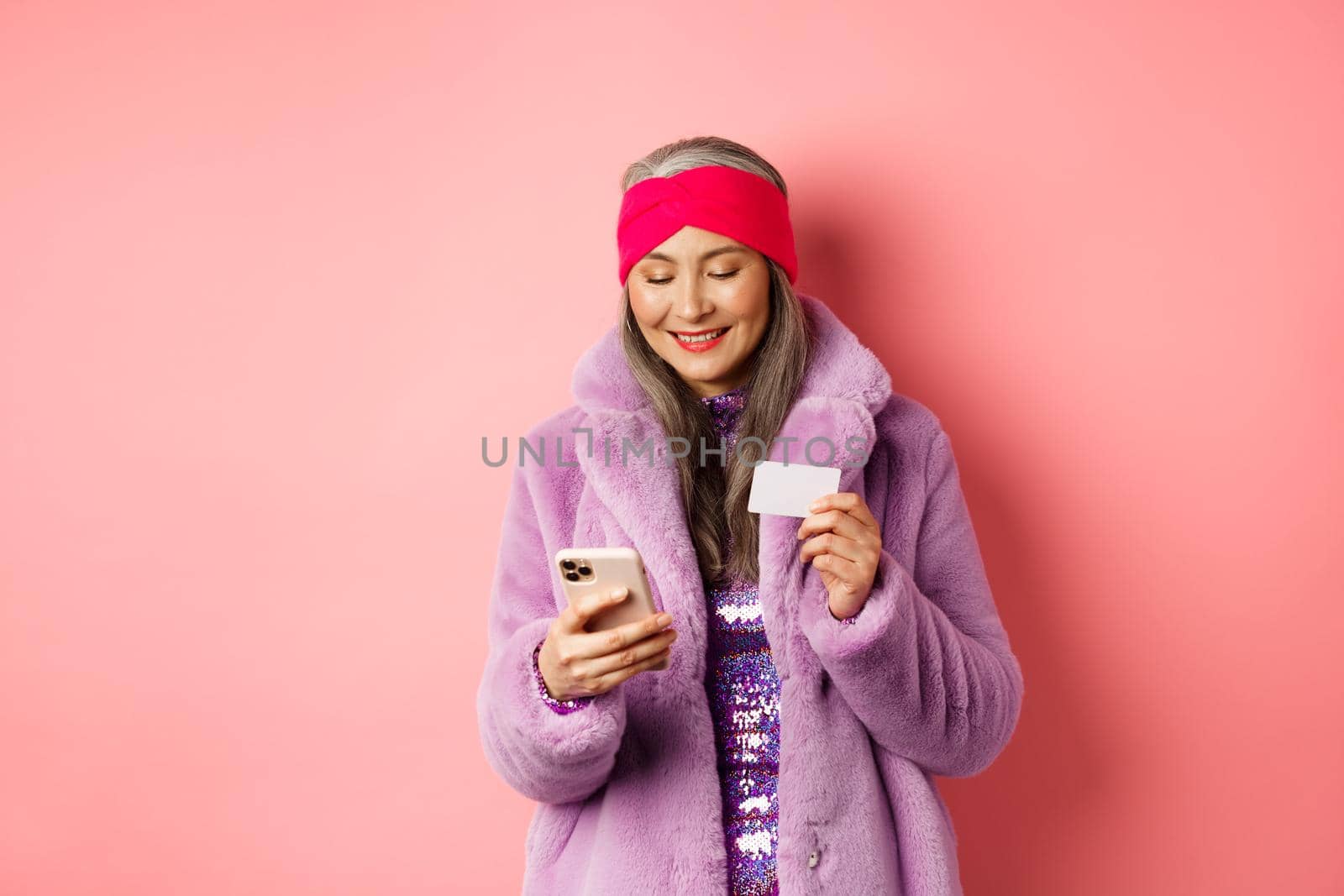 Online shopping and fashion concept. Stylish asian elderly woman standing in fashionable purple coat and making payment on smartphone, holding plastic credit card, pink background.