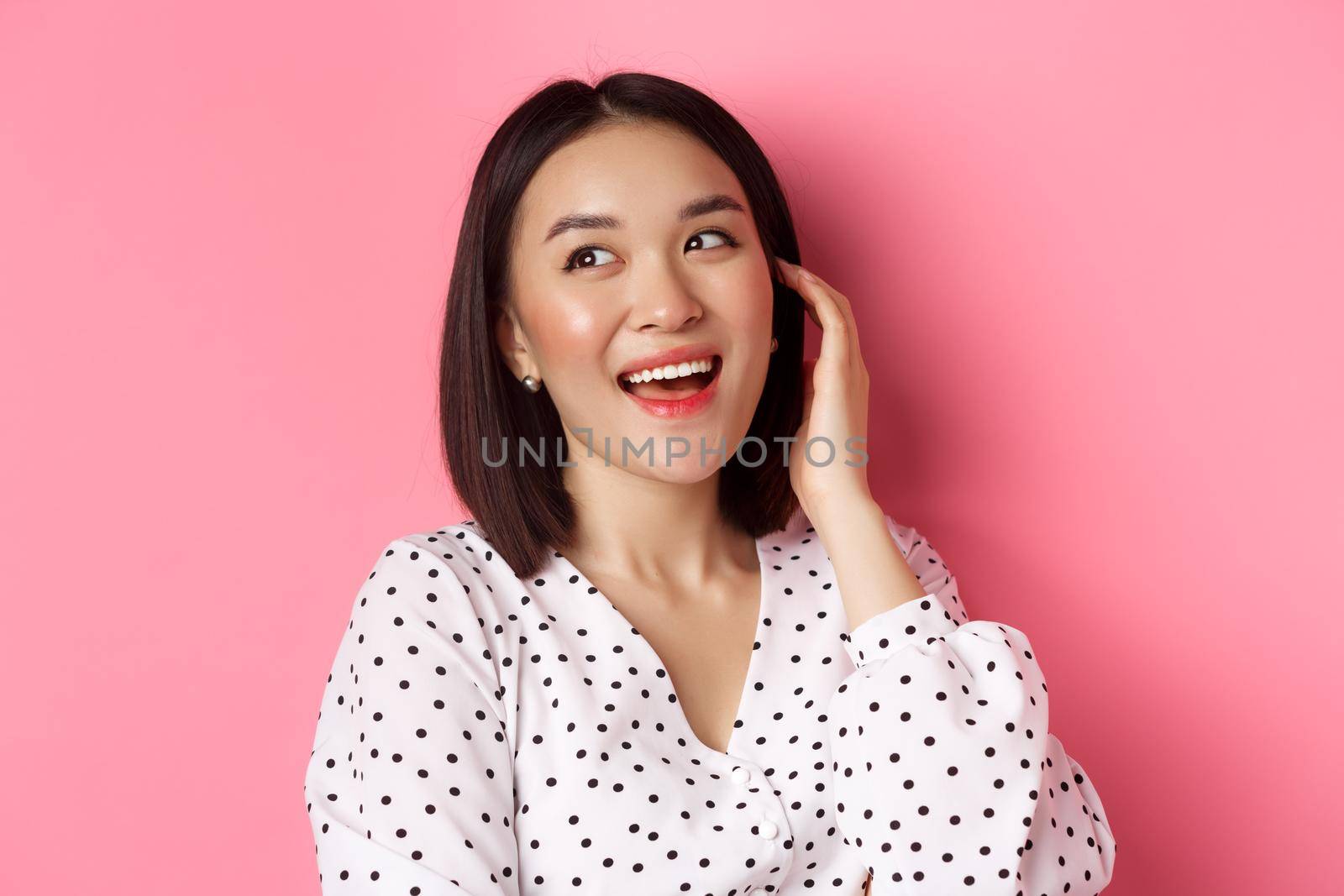 Close-up of flirty asian woman sighing, looking dreamy left and smiling, standing over pink background. Copy space