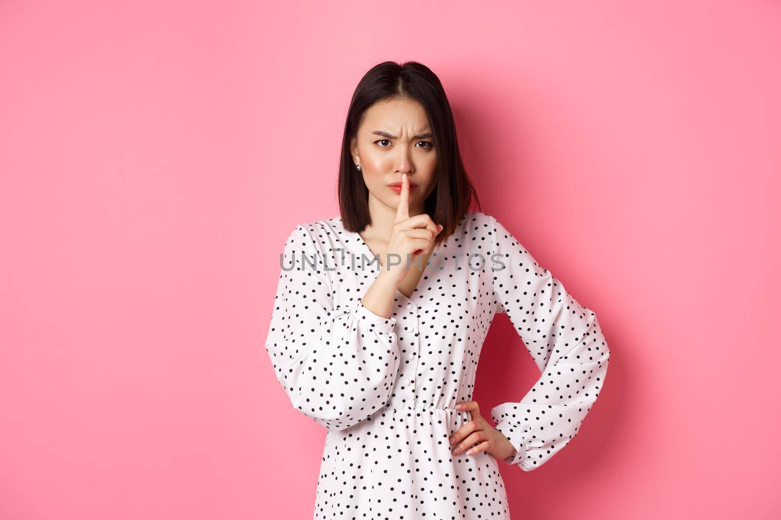 Angry asian female model shushing, frowning and hushing, showing taboo gesture, standing displeased against pink background, demand silence.
