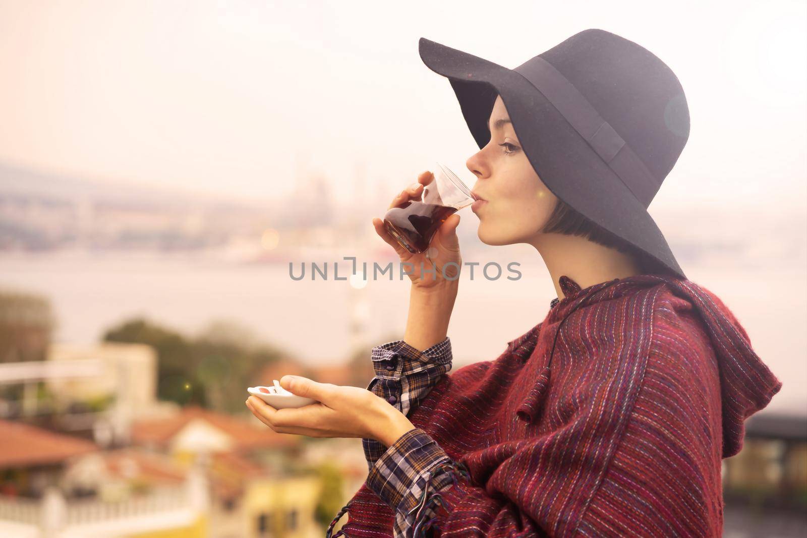 A young girl in a poncho and a hat drinks fragrant delicious tea on the background of the ocean while traveling.