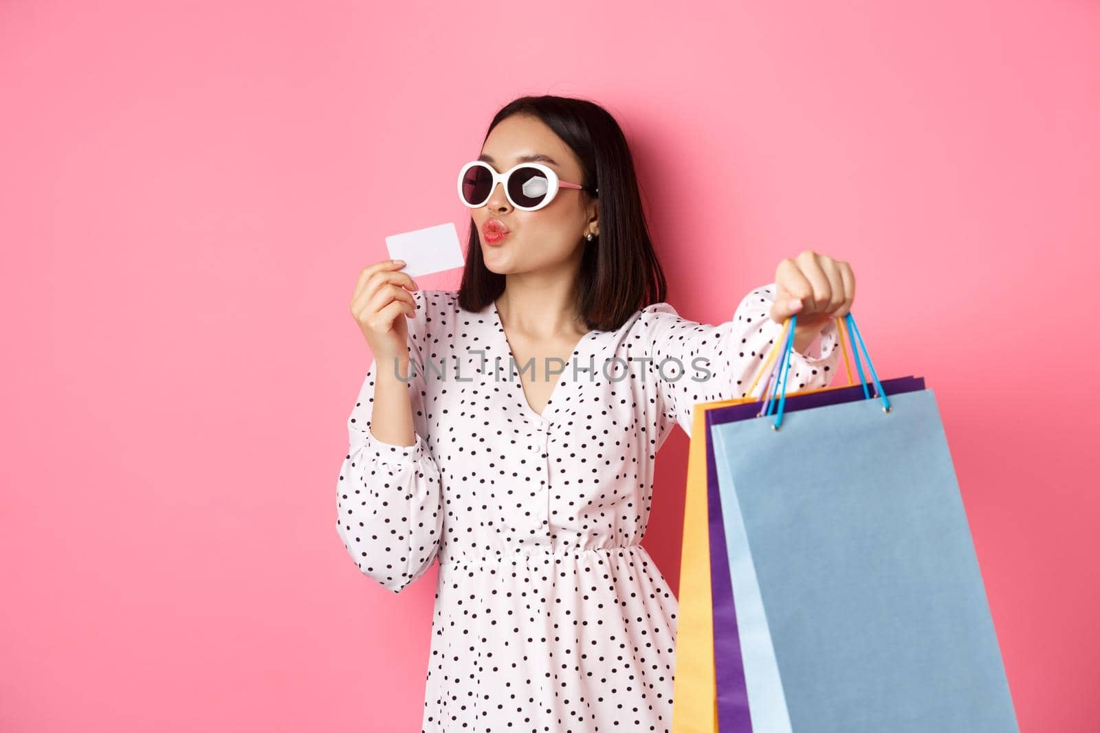 Beautiful asian woman in sunglasses going shopping, holding bags and kissing credit card, standing over pink background.