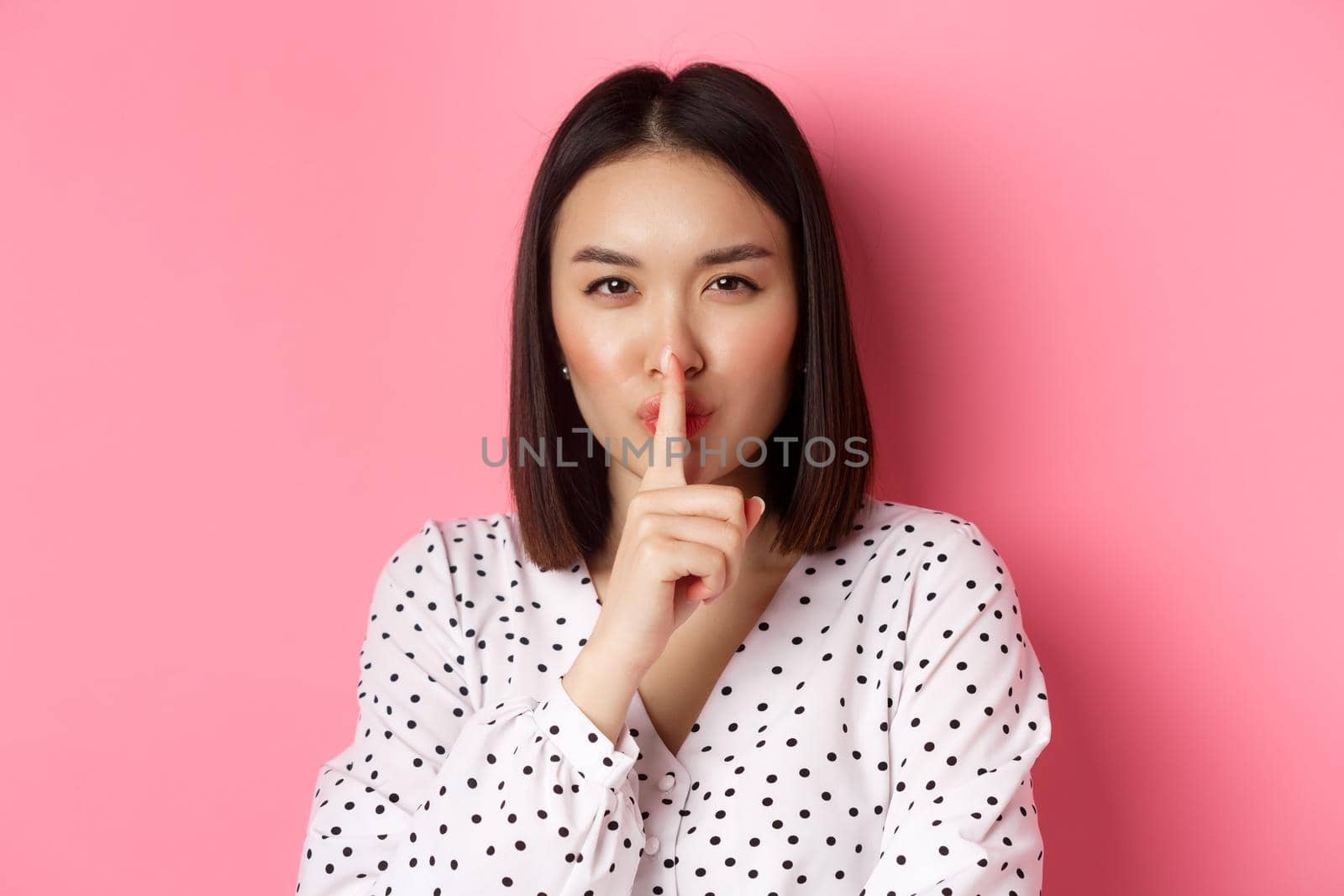 Close-up of mysterious asian woman hiding a secret, hushing and telling to keep quiet, standing over pink background. Copy space