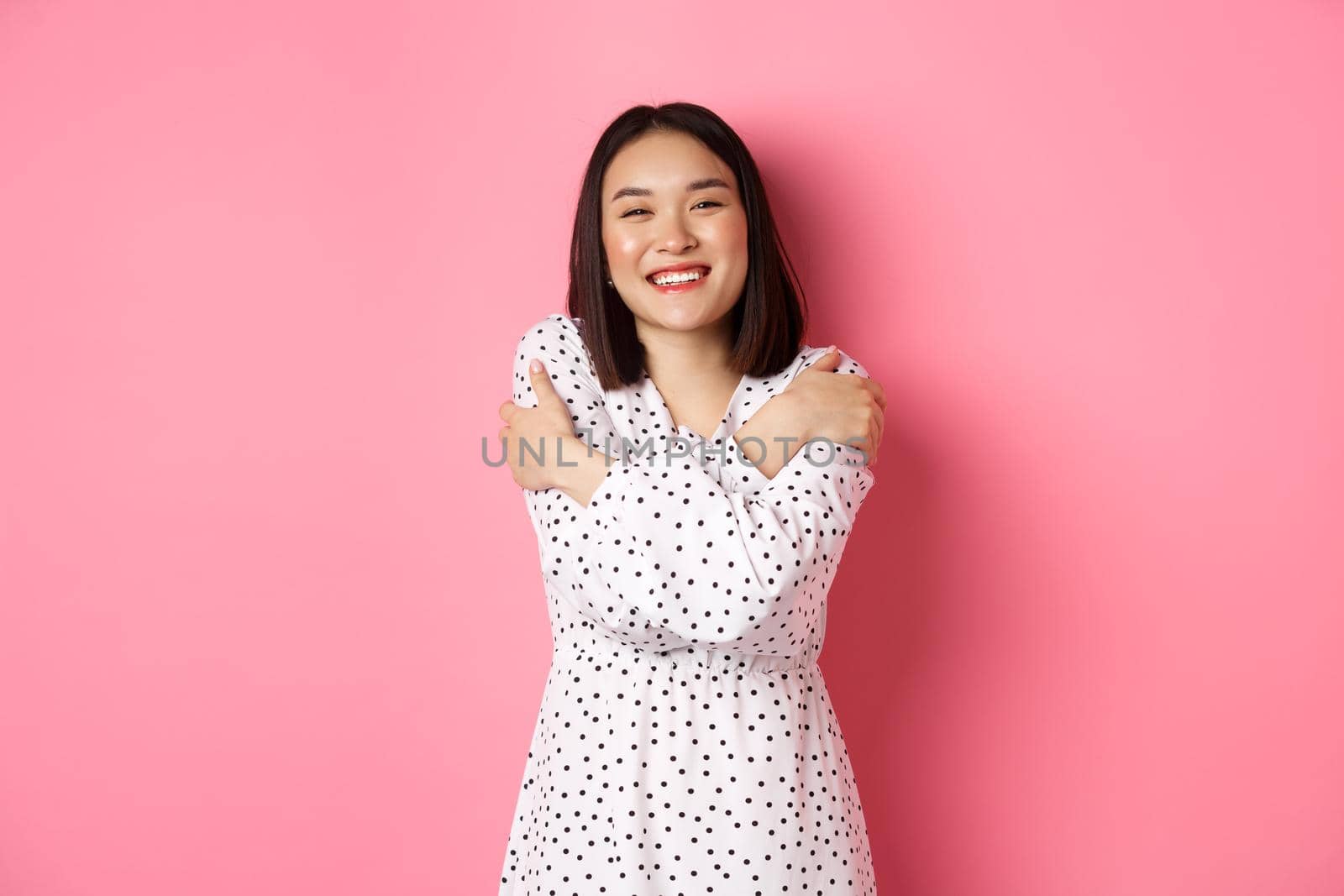 Beautiful asian girl embracing own body, hugging herself and smiling with happy face, looking at camera carefree, standing over pink background.