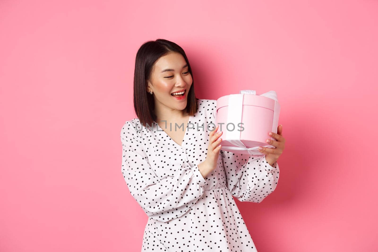 Valentines day, celebration concept. Beautiful asian woman holding romantic gift box, smiling happy, standing over pink background.