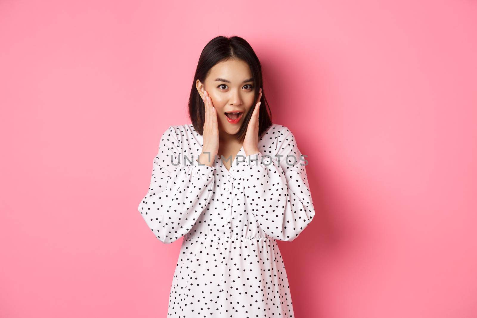 Young asian woman looking intrigued, gossiping and saying wow with curious face, looking interested at camera, standing over pink background.