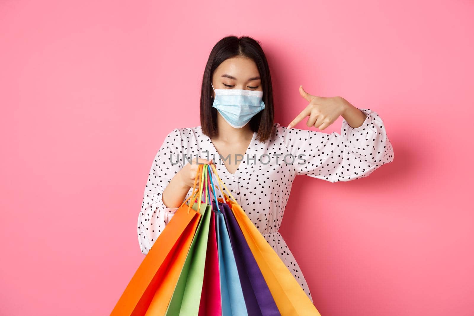 Covid-19, pandemic and lifestyle concept. Pretty korean woman in medical mask pointing at shopping bags, buying clothes, standing over pink background.