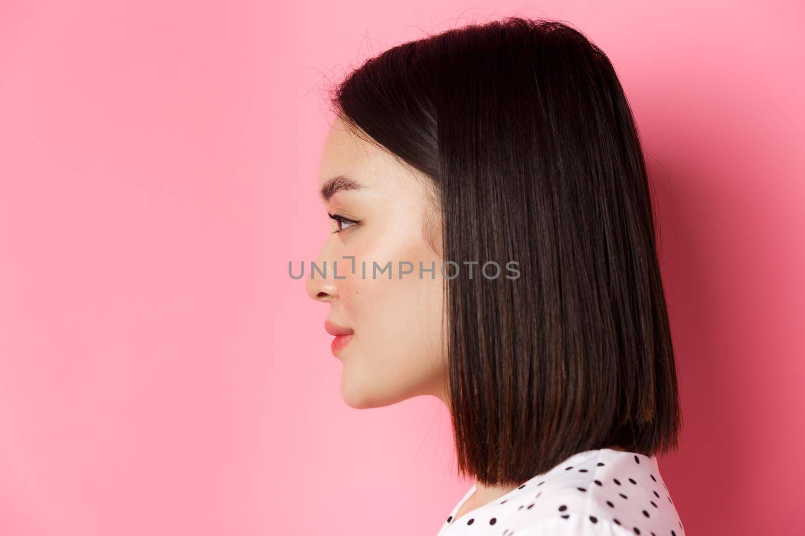 Beauty and skin care concept. Headshot profile of young beautiful asian woman with short dark hair, looking left at copy space, standing over pink background.