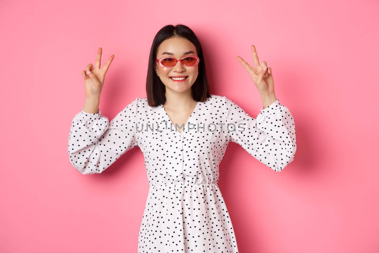 Tourism and lifestyle concept. Beautiful asian woman on summer vacation, showing peace signs and smiling happy, wearing trendy sunglasses, pink background.