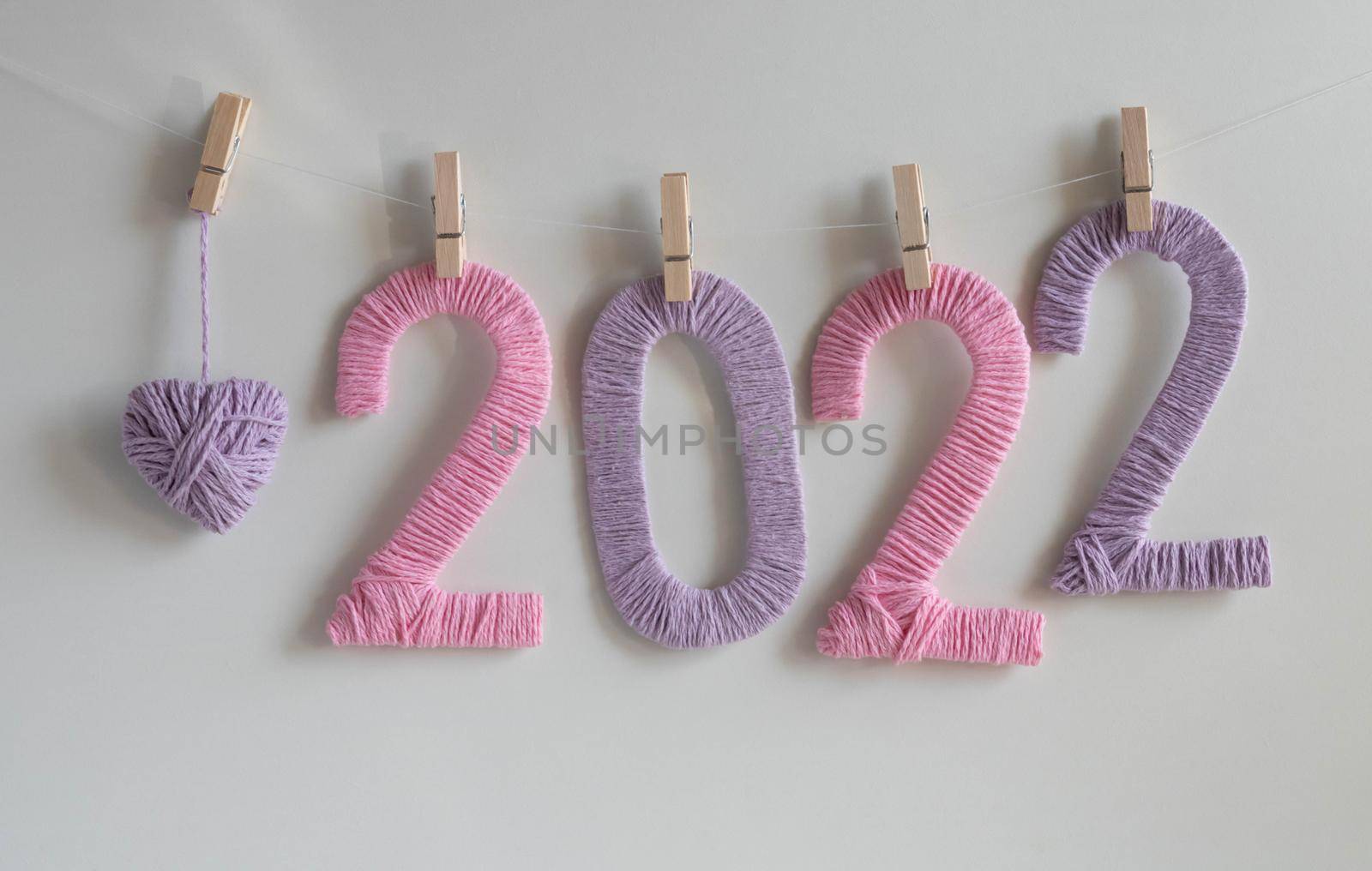The numbers 2022, made of pink and lilac threads on a white background, hang on clothespins next to a lilac knitted heart. The concept of the New Year.