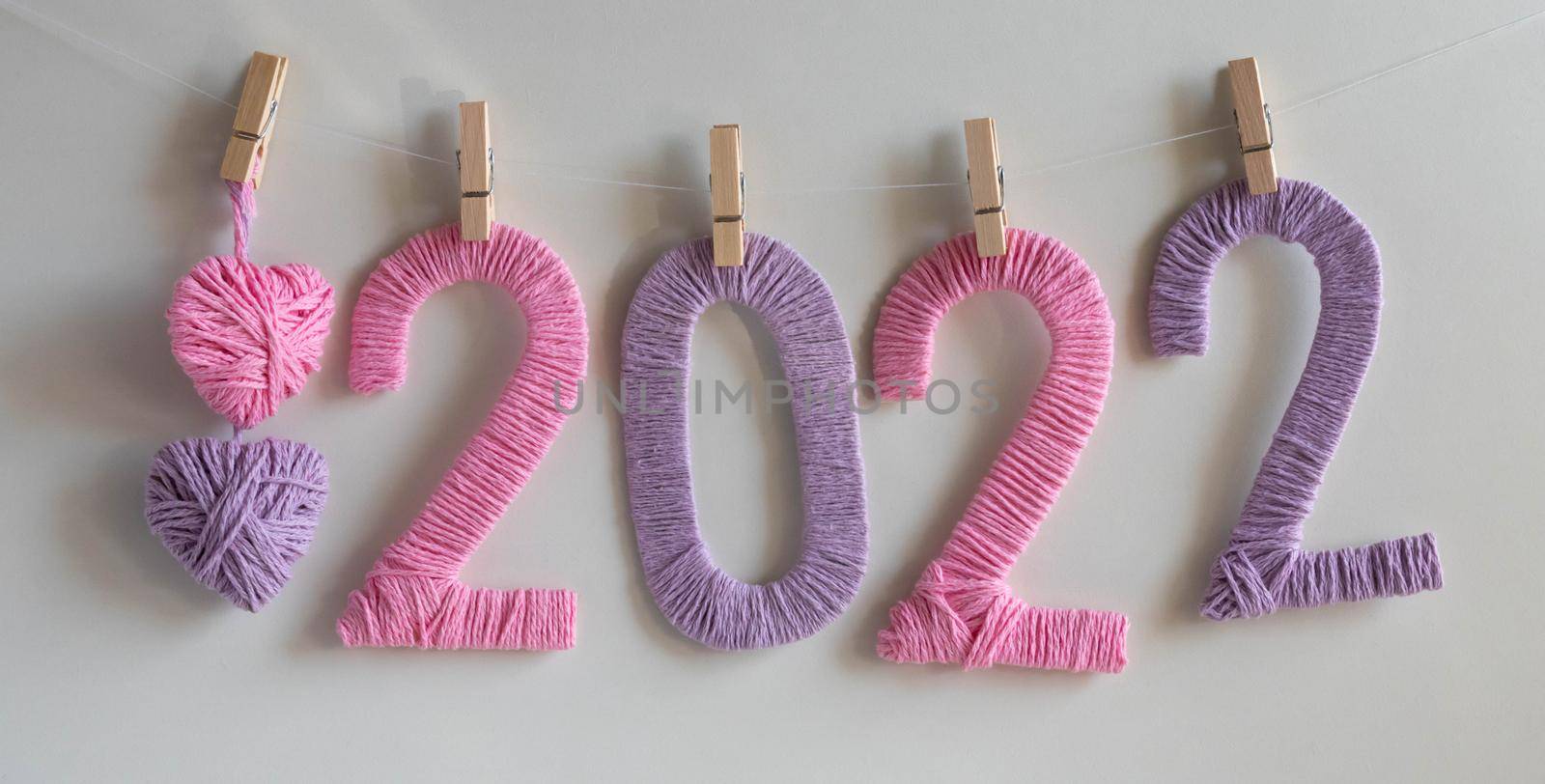 The numbers 2022, made of pink and lilac threads on a white background, hang on clothespins next to lilac and pink knitted hearts. The concept of the New Year by lapushka62