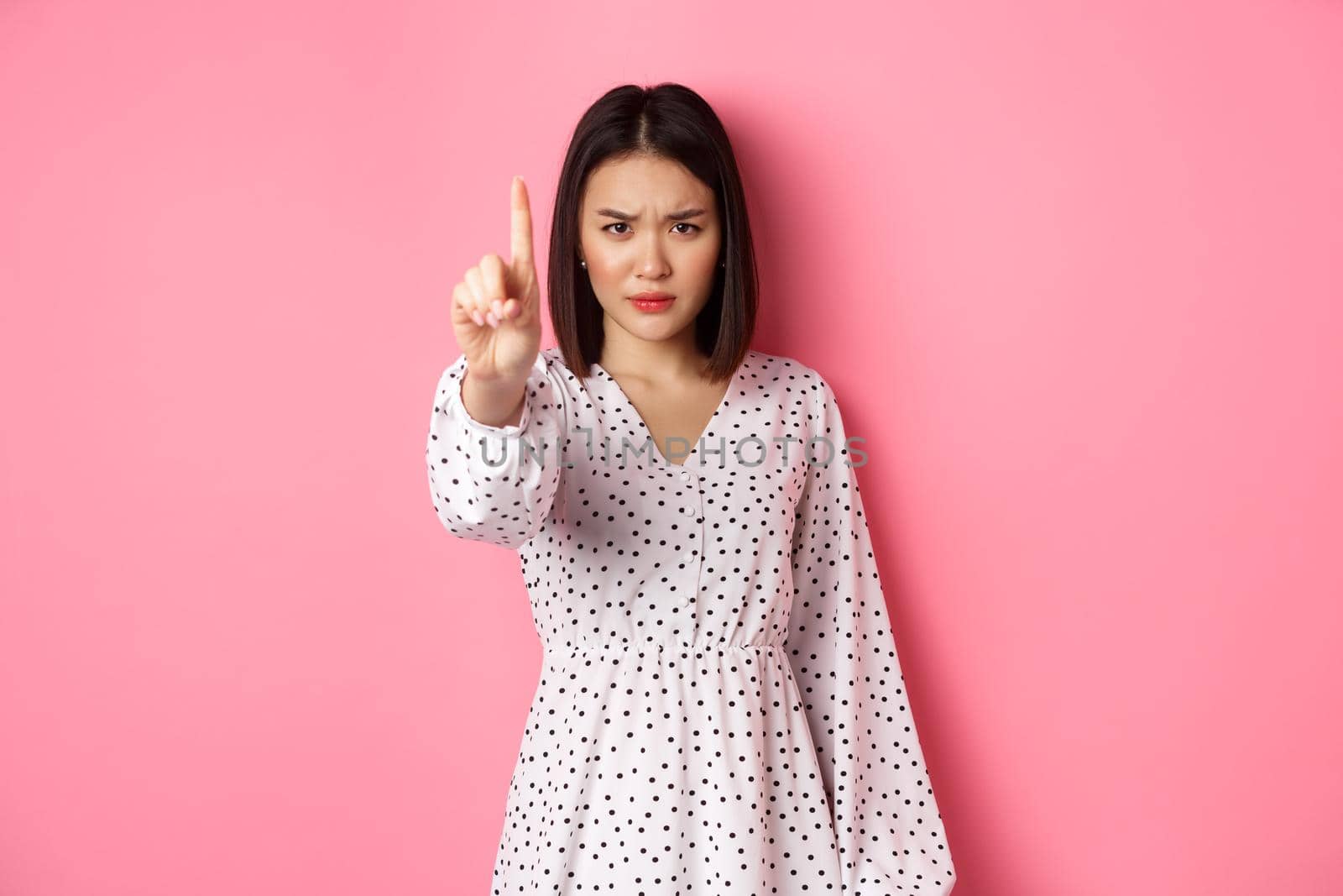 Serious and angry asian woman telling to stop, frowning and showing finger in disapproval, prohibit something, standing in dress against pink background.