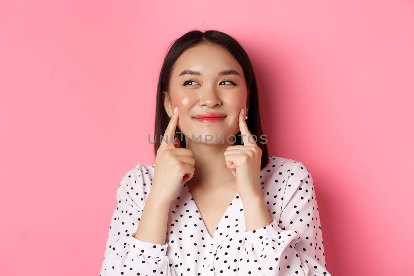 Beauty and skin care concept. Close-up of beautiful asian woman poking cheeks and smiling satisfied, standing over pink background.