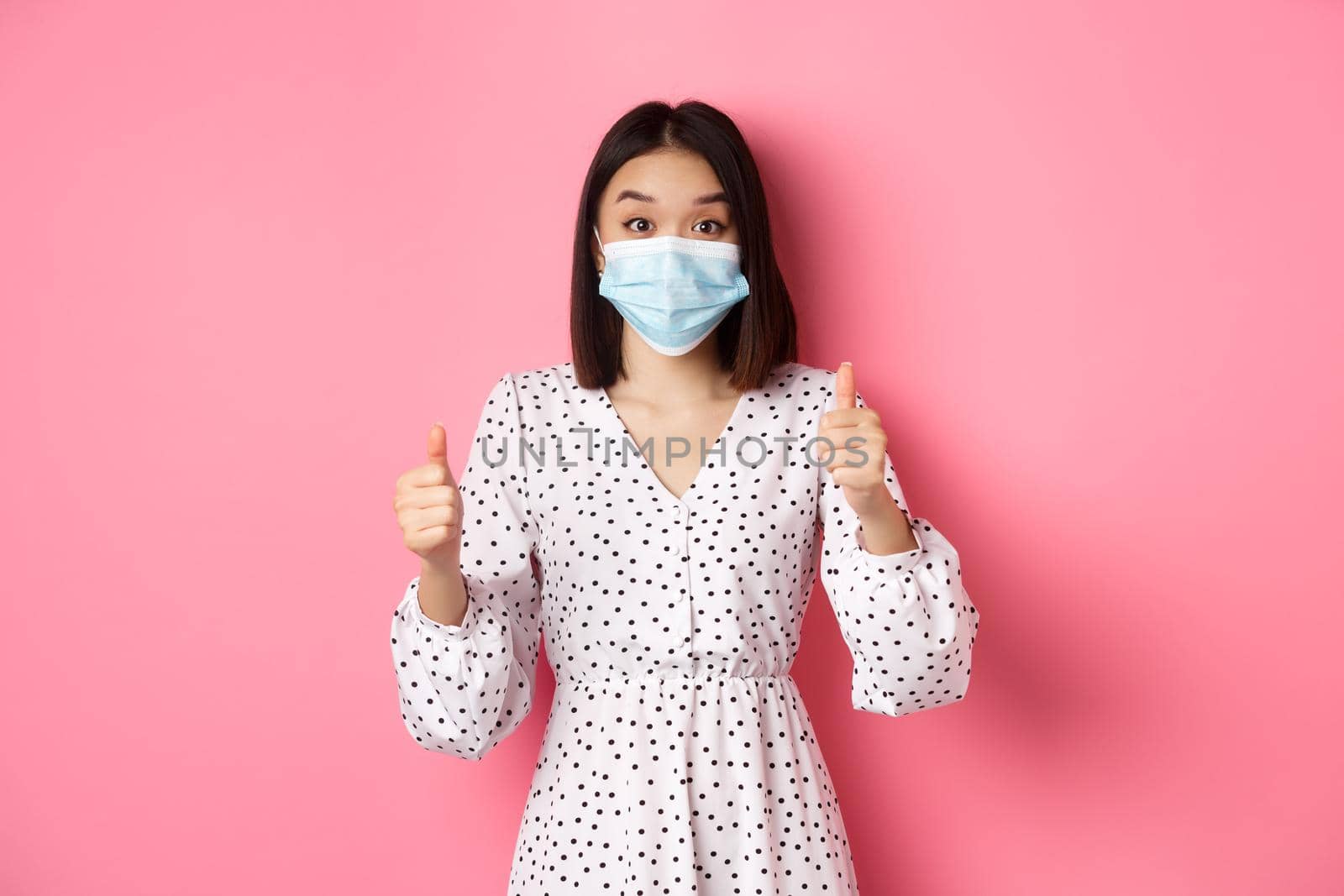 Covid-19, quarantine and lifestyle concept. Excited asian woman in face mask showing thumbs-up, praising good job, looking amazed at camera, standing over pink background.