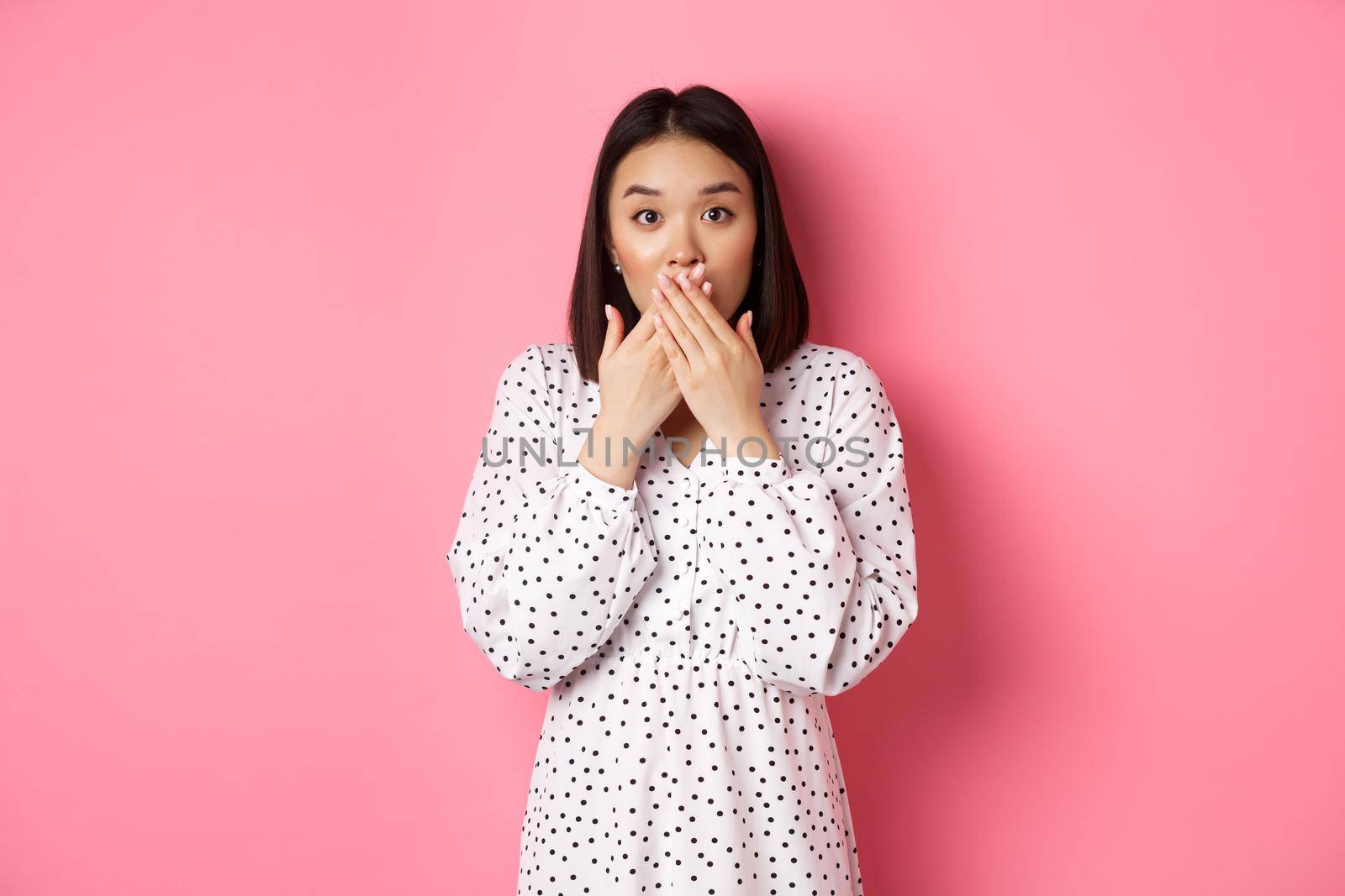 Image of surprised asian female model gasping, cover mouth and looking at camera, standing amazed against pink background.
