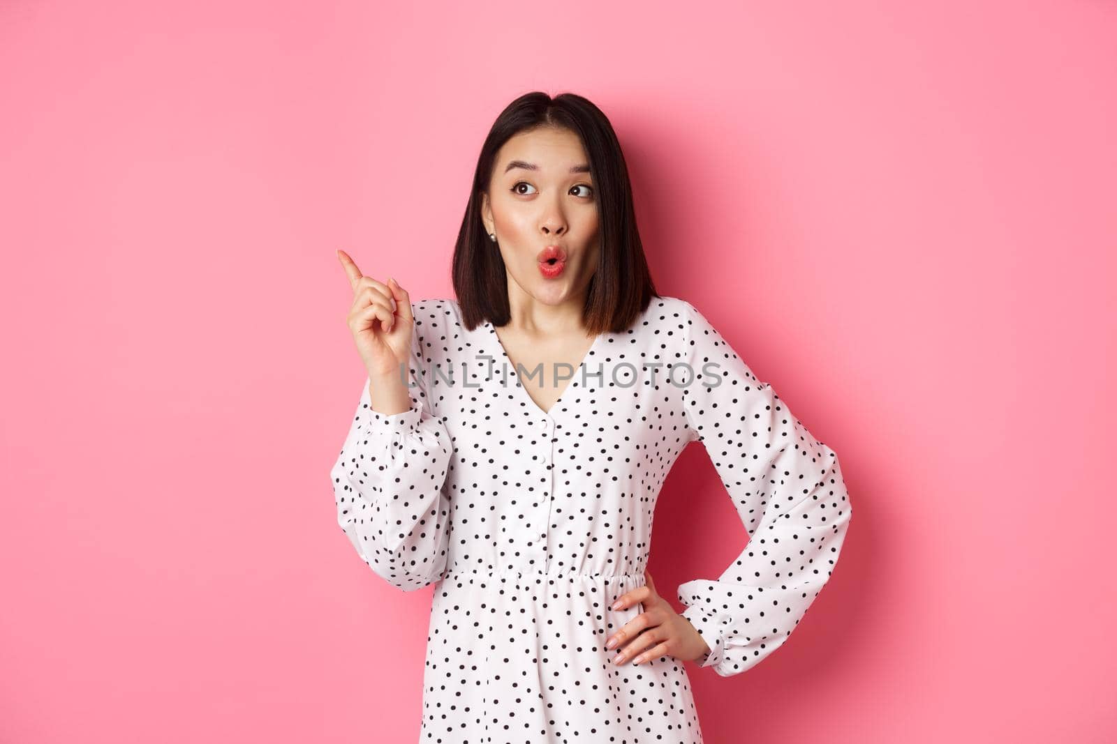 Amazed asian girl in dress pointing finger at upper left corner promo offer, looking interested, shopping and standing over pink background.