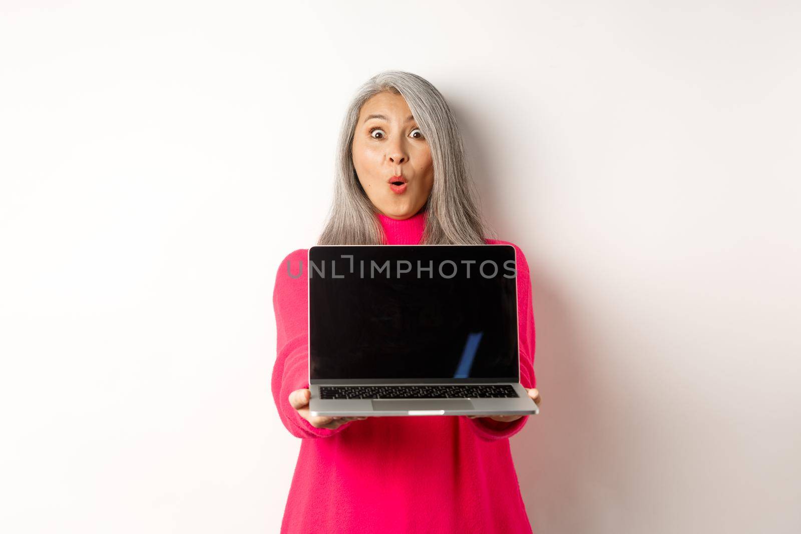 E-commerce concept. Impressed asian senior woman showing blank laptop screen and staring amazed at camera, standing over white background.