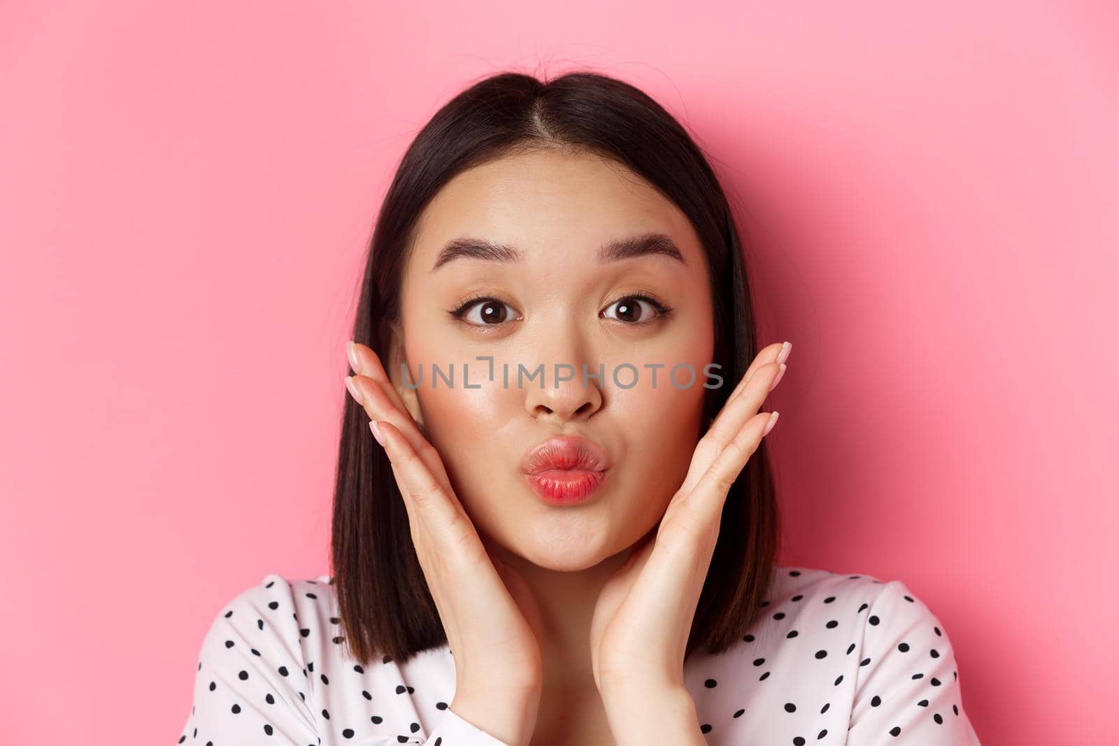 Beauty and lifestyle concept. Close-up of adorable asian woman touching face, pucker lips in kiss, standing over pink background.