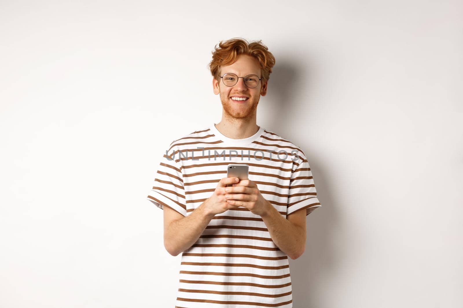 Technology and e-commerce concept. Redhead guy in glasses using mobile phone and smiling. Young man with smartphone staring happy at camera, white background.