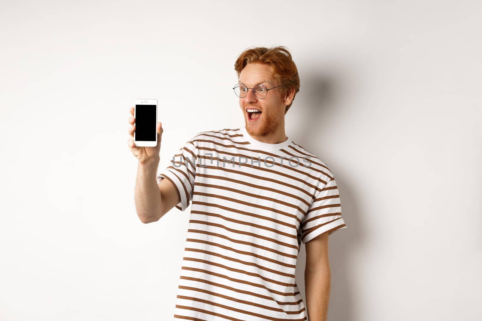 Technology and e-commerce concept. Happy young redhead man in glasses showing blank smartphone screen, looking amazed, standing over white background.