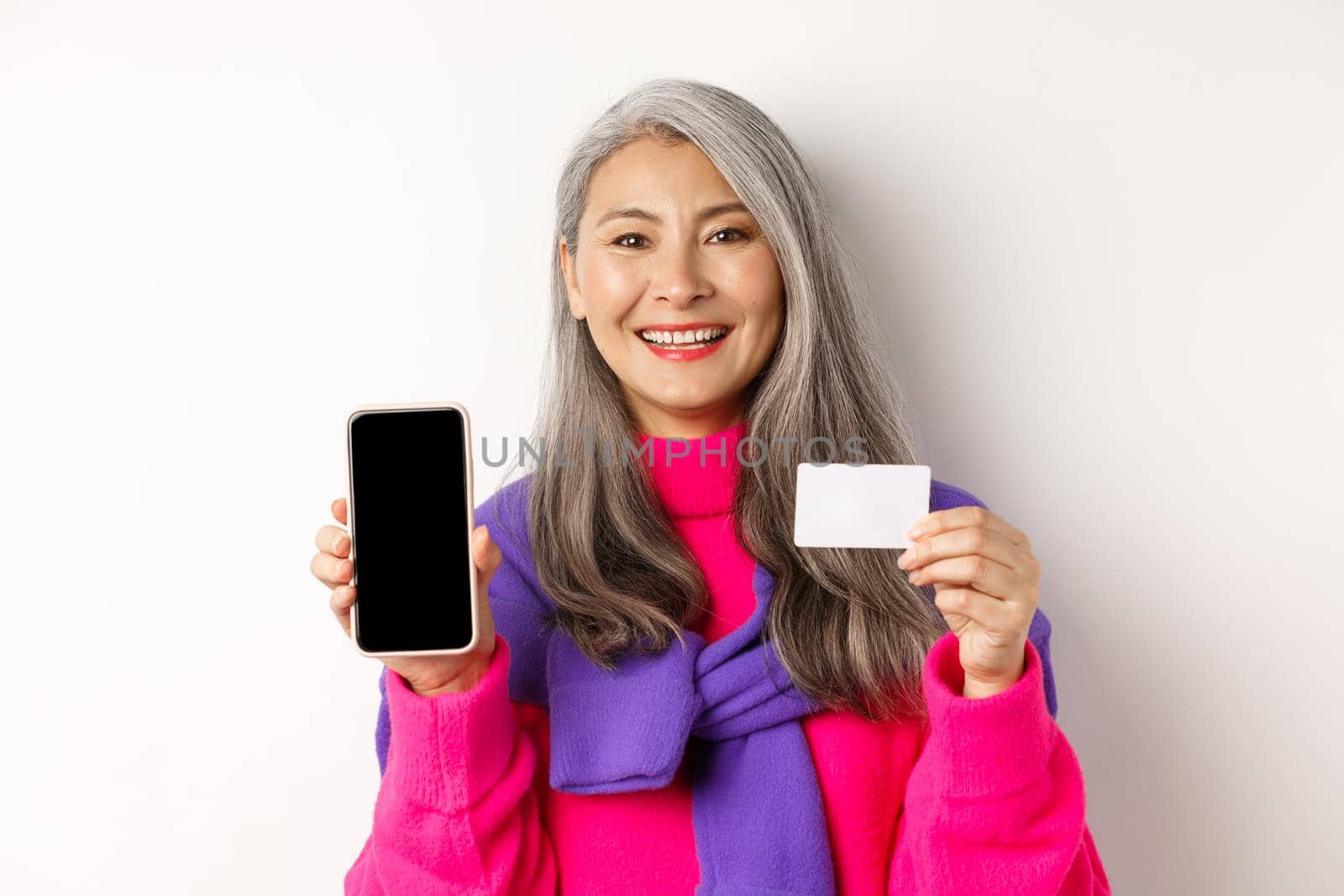 Online shopping. Closeup of smiling asian senior woman showing blank mobile screen and plastic credit card, looking happy, standing over white background.