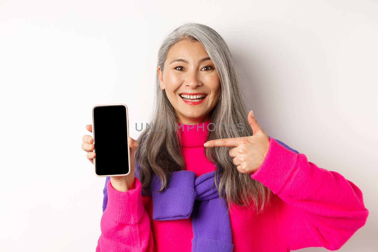 Online shopping. Beautiful asian grandmother smiling, pointing finger at smartphone blank screen, showing mobile application, standing over white background.