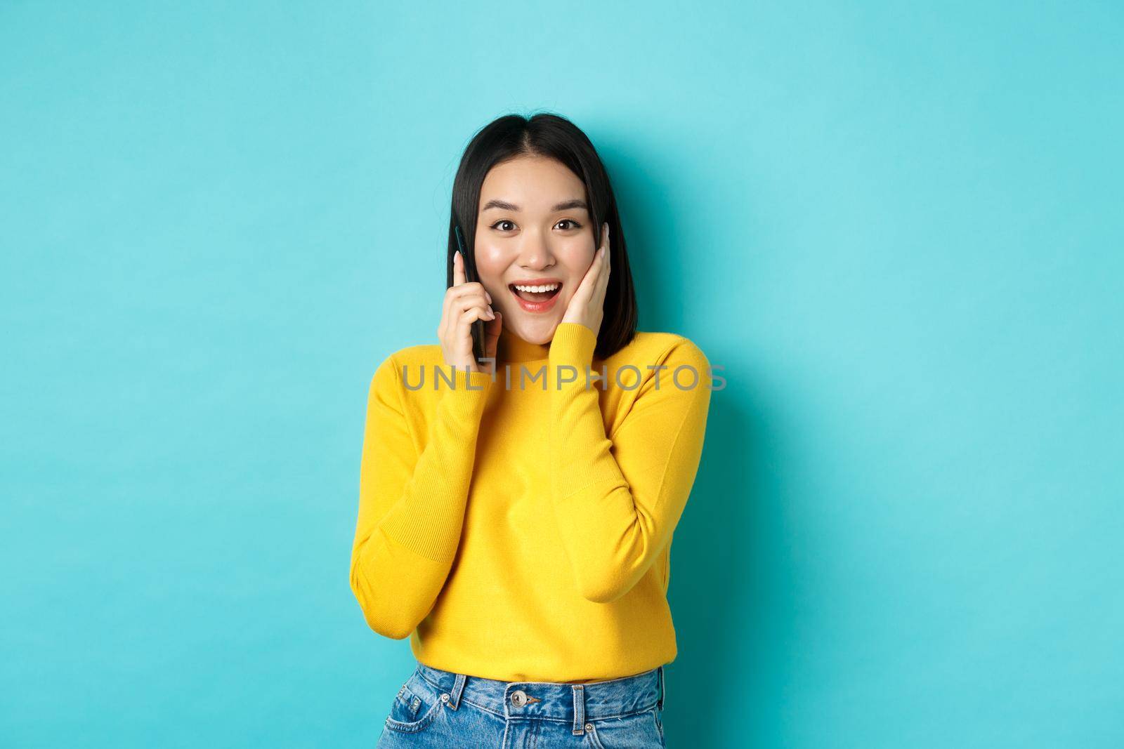 Happy asian woman receive offer on phone call, smiling while talking on smartphone, standing over blue background.