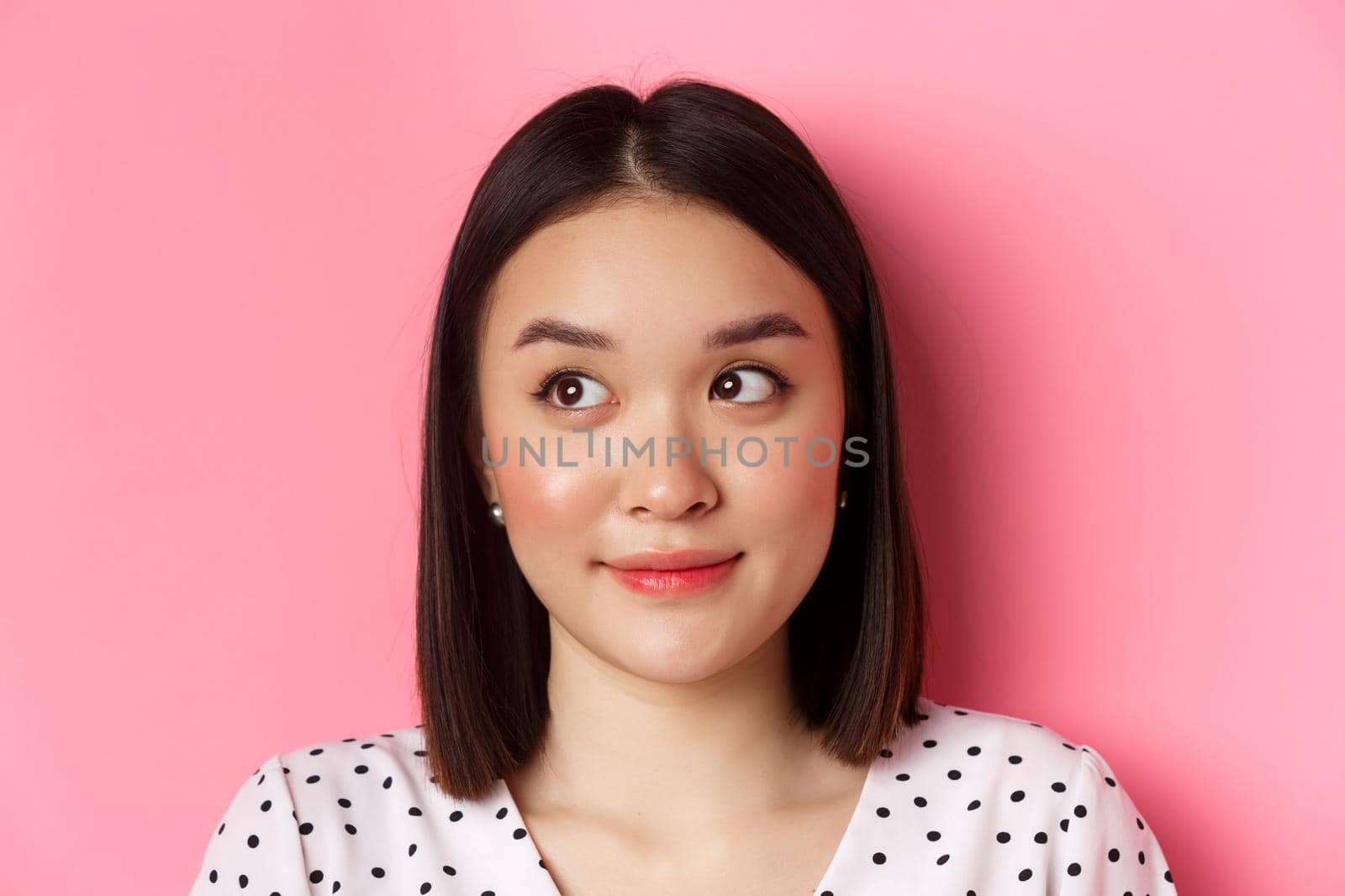 Beauty and skin care concept. Close-up of cute asian teenage girl looking left at banner, smiling silly, standing in dress over pink background.