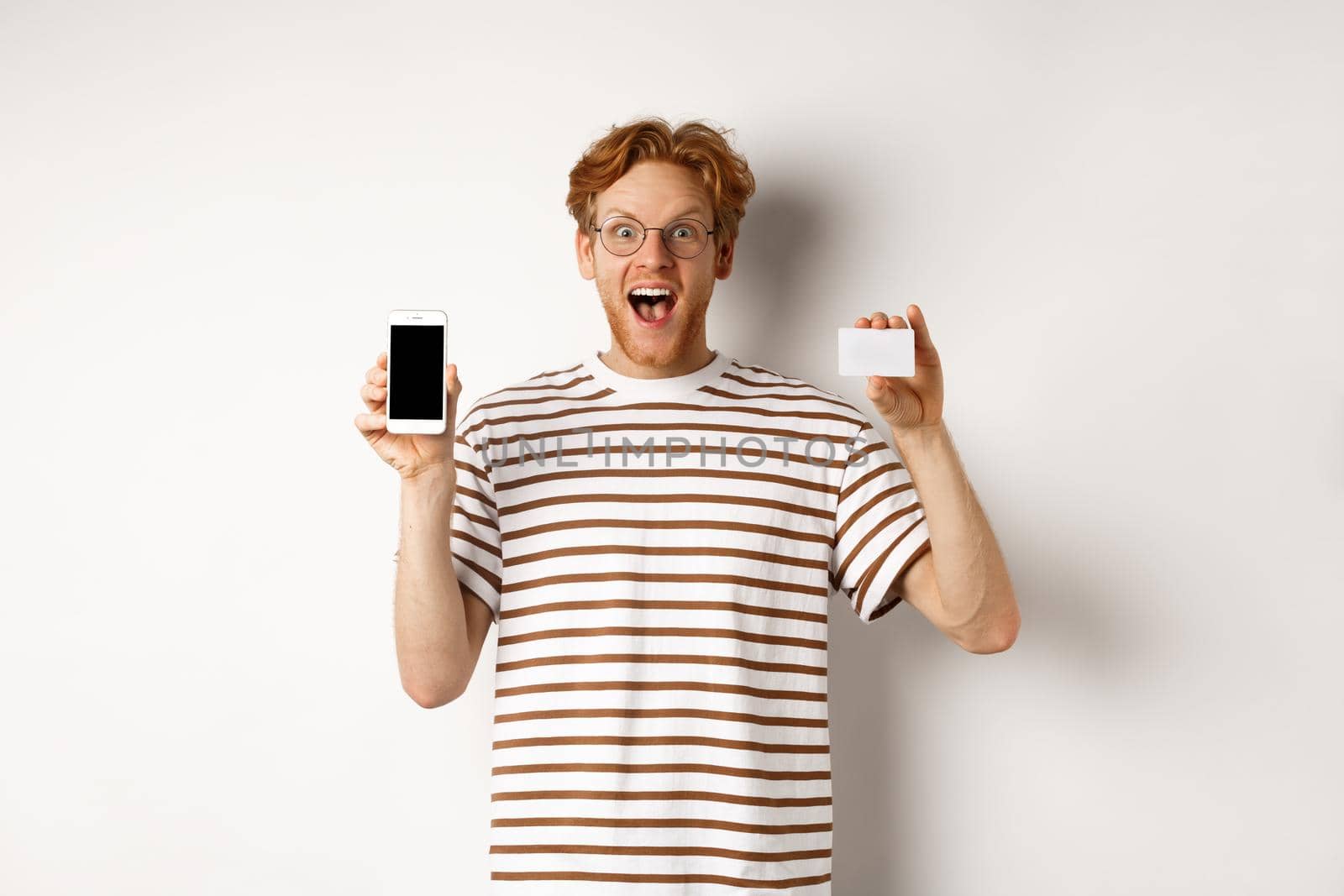 Shopping and finance concept. Young man showing blank mobile screen and plastic credit card, smiling at camera, white background.