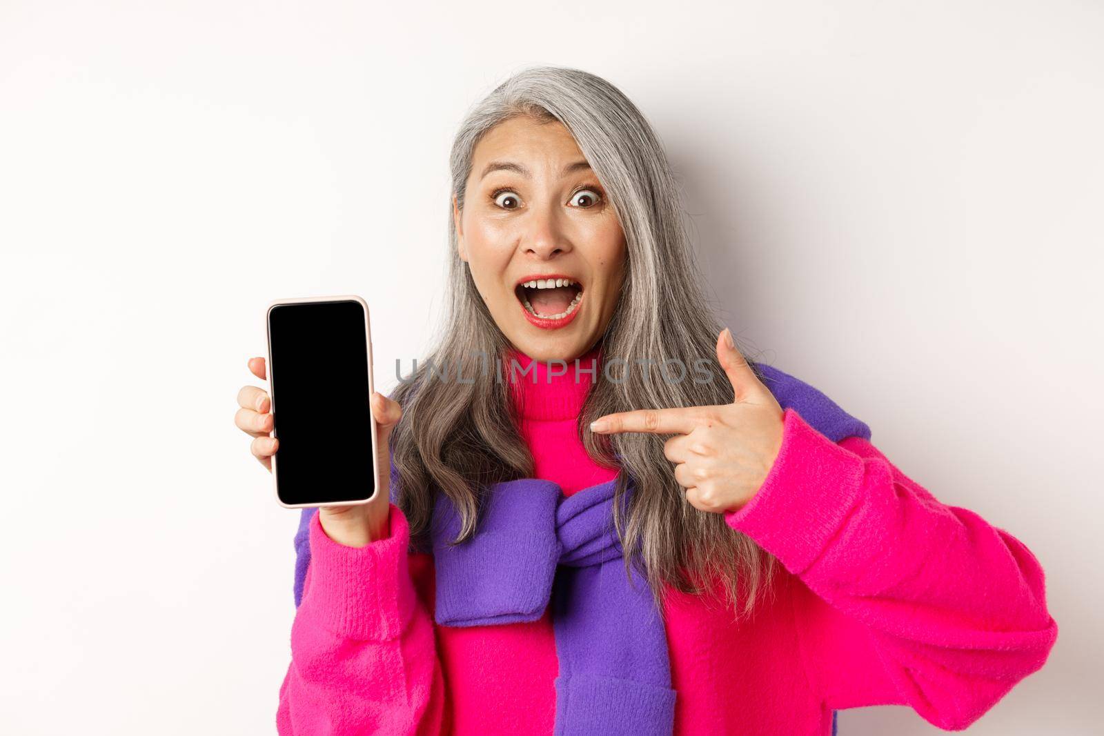 Online shopping. Beautiful asian grandmother smiling, pointing finger at smartphone blank screen, looking amazed, showing mobile application, standing over white background.