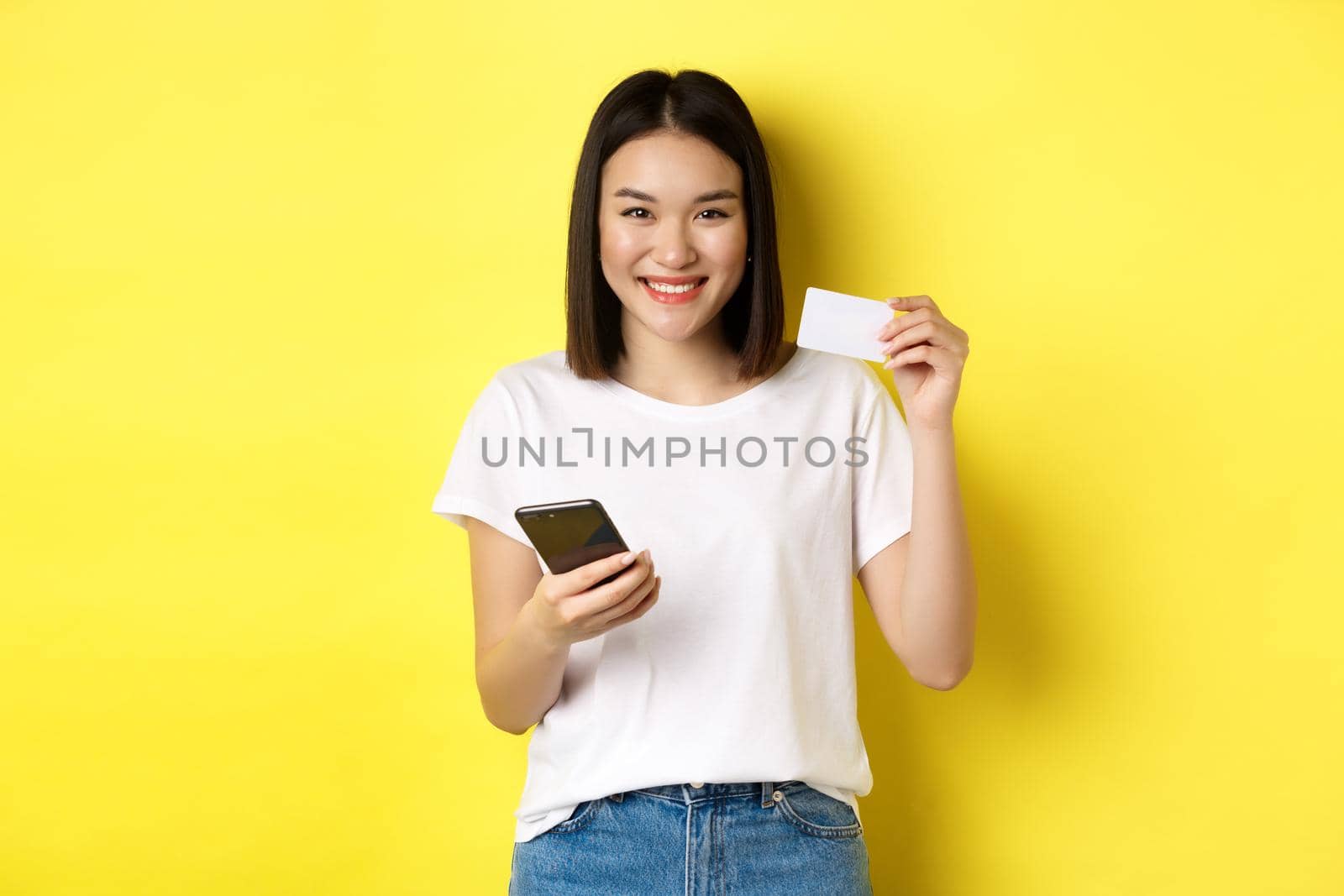 Attractive korean woman paying online with smartphone, showing plastic credit card and smiling, standing over yellow background.