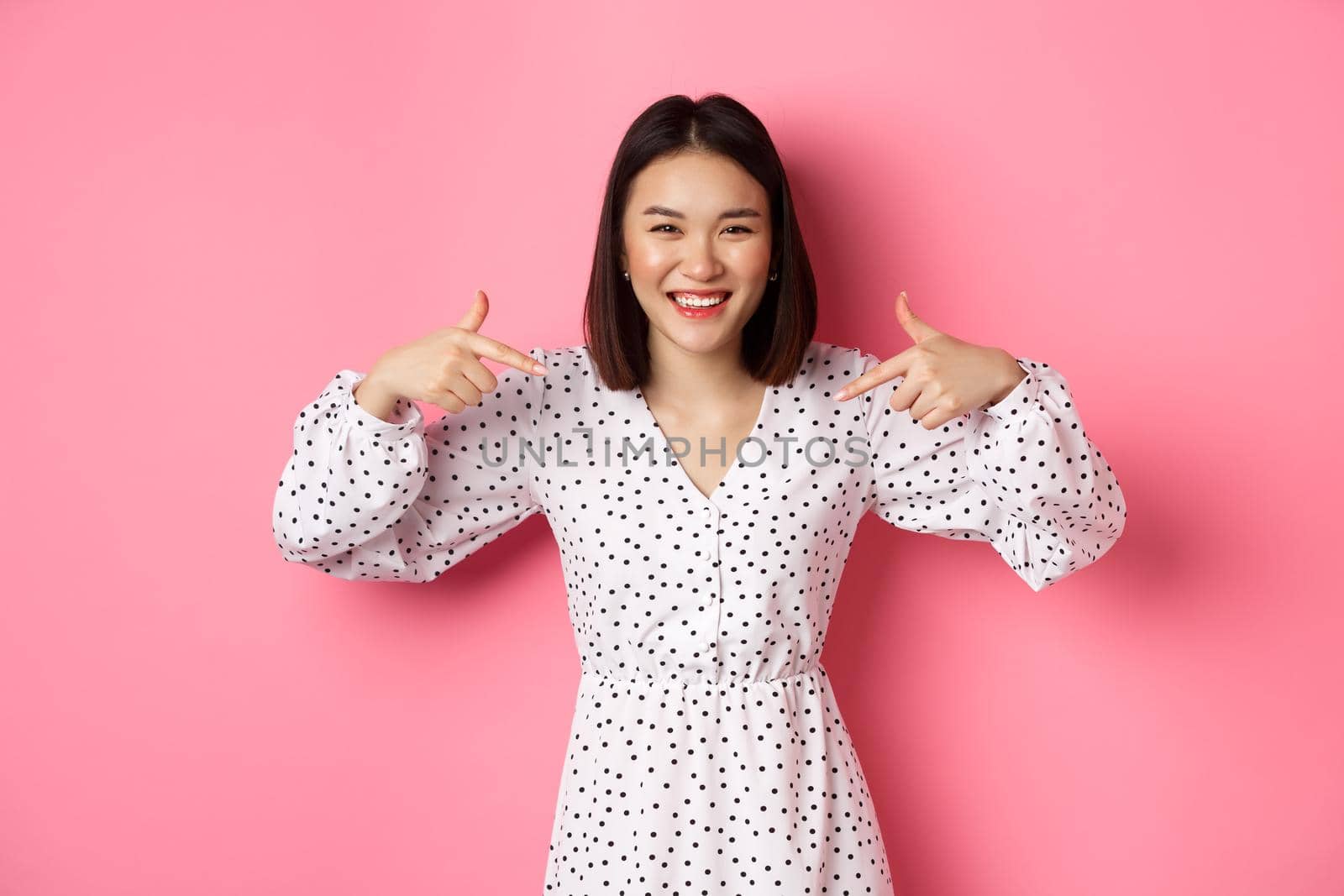 Beautiful korean woman pointing fingers at your logo and smiling, standing in dress over romantic pink background. Copy space