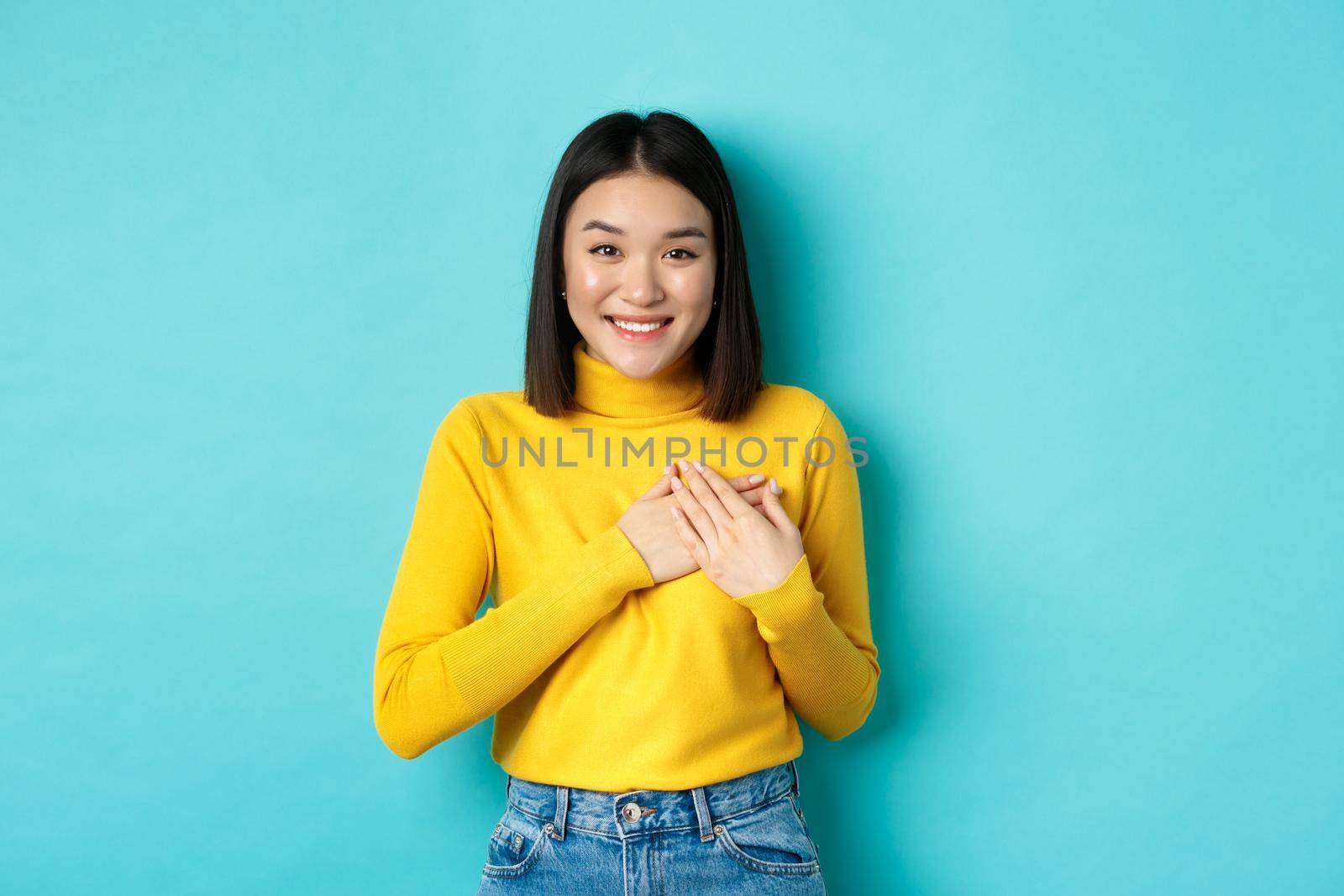 Image of beautiful asian woman holding hands on heart and smiling, thanking you, feeling grateful, standing over blue background.