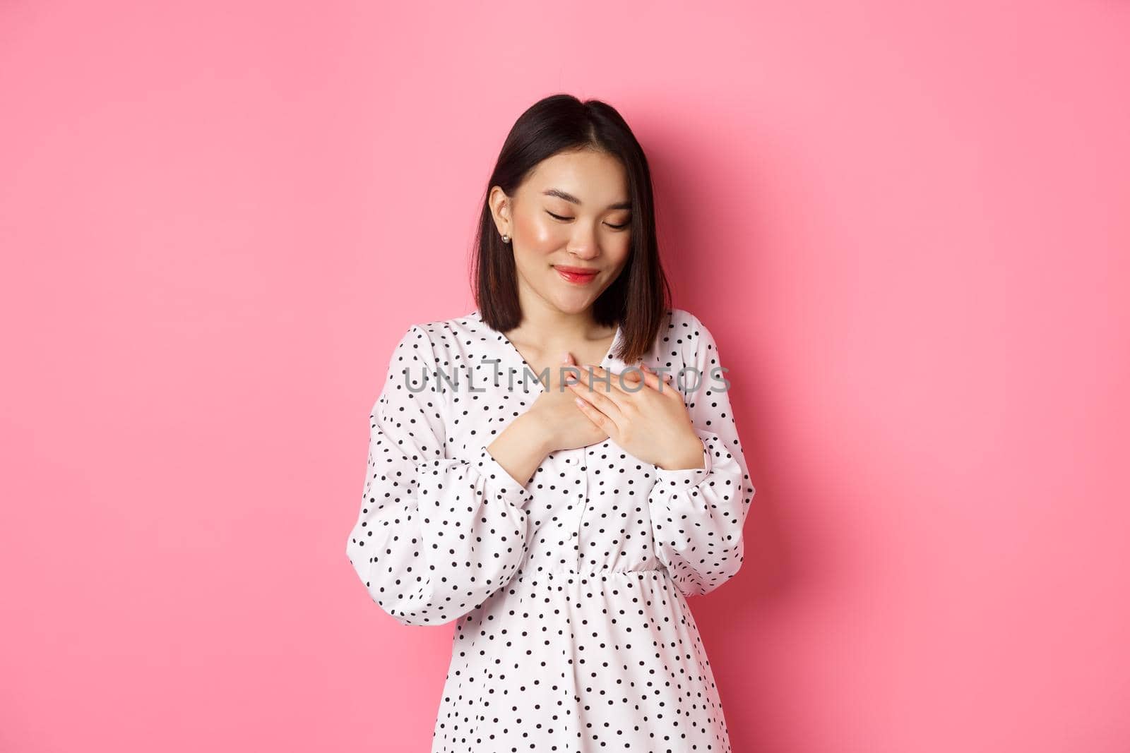 Beautiful korean girl in trendy spring dress dreaming, holding hands on heart, smiling with closed eyes, imaging something or having heartwarming memory, pink background.