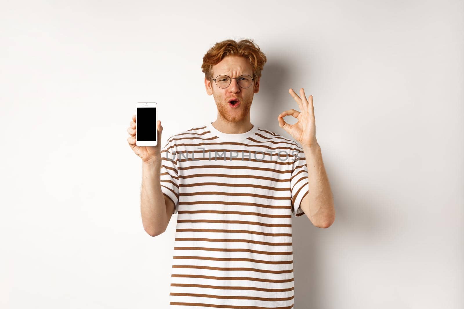 Technology and e-commerce concept. Young man with red hair showing okay sign and blank smartphone screen, praising awesome app or video game, white background.