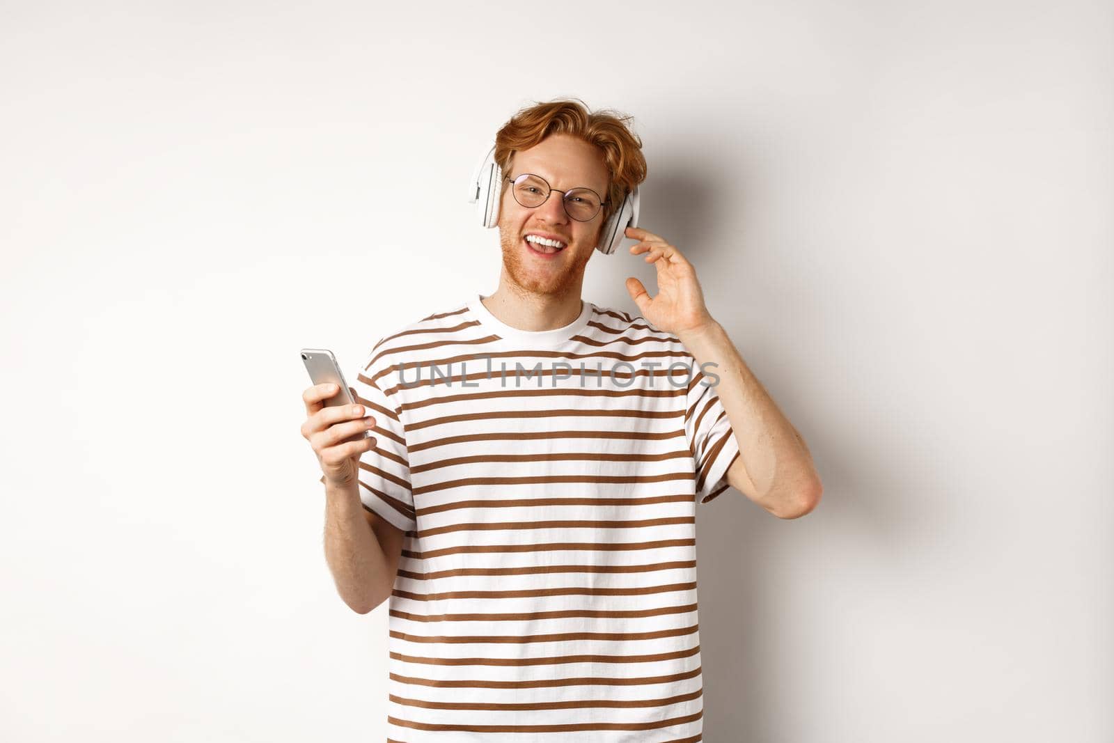 Technology concept. Young man with red hair and beard listening music in headphones and using smartphone, smiling at camera, white background by Benzoix