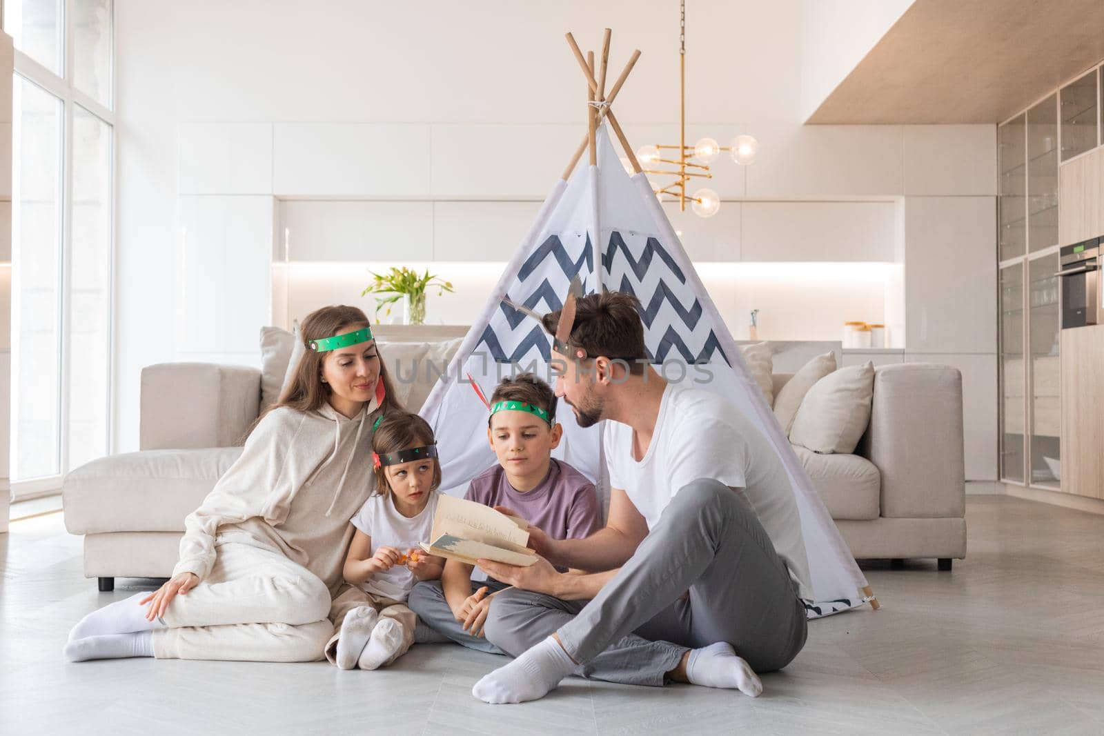 Happy family of parents and two children playing indian at home, wigwam tent, feather roach, reading book about native american indian culture