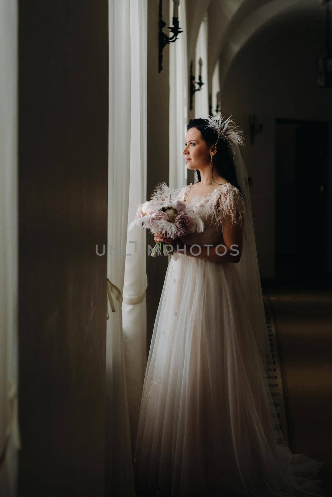 The bride in a wedding dress and with a bouquet stands at the old window and looks.