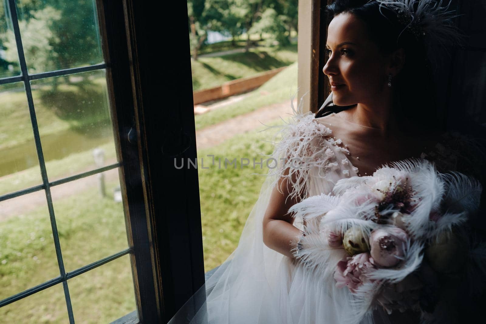 A bride in a wedding dress and a bouquet sits at an open old window and looks.